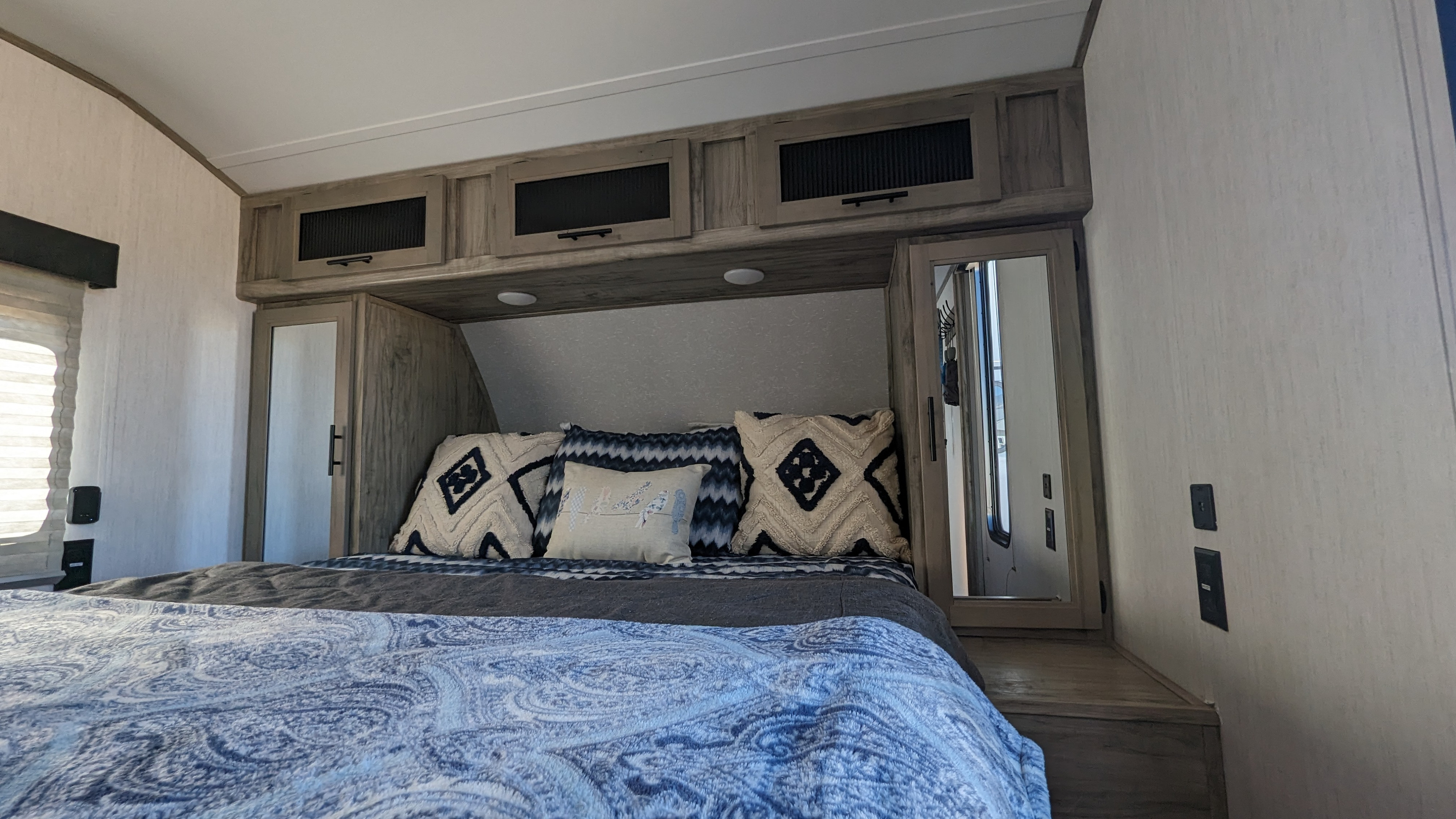 Cozy RV rental bedroom featuring a patterned bedspread, decorative pillows, and compact overhead storage with mirrored cabinets.. Forest River Cherokee Alpha Wolf 2021