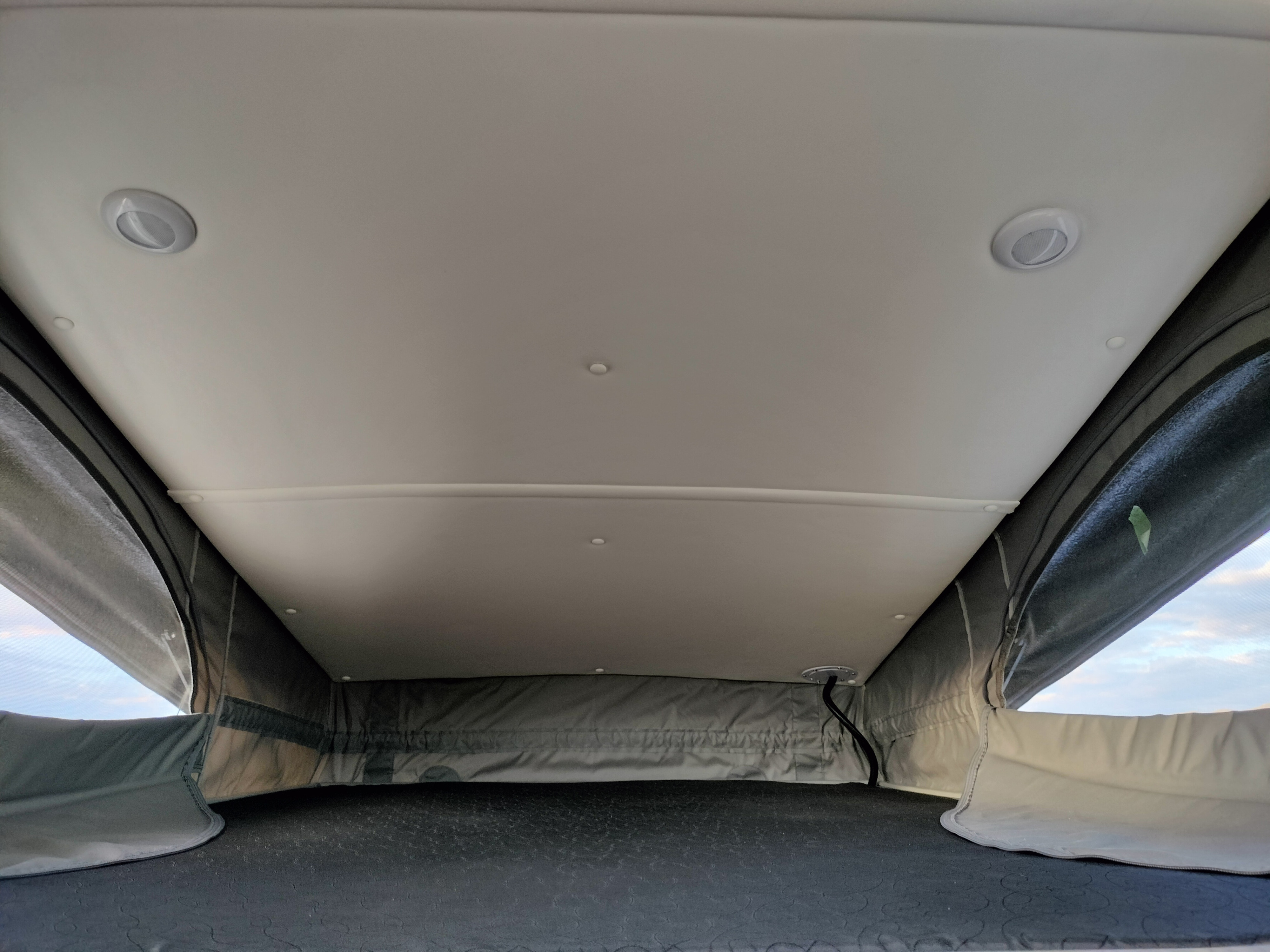 An RV rental's pop-top interior: ceiling lights illuminate the bed, surrounded by fabric walls showing outside views.. Ram ProMaster 1500 Tofino Camper Van 2023