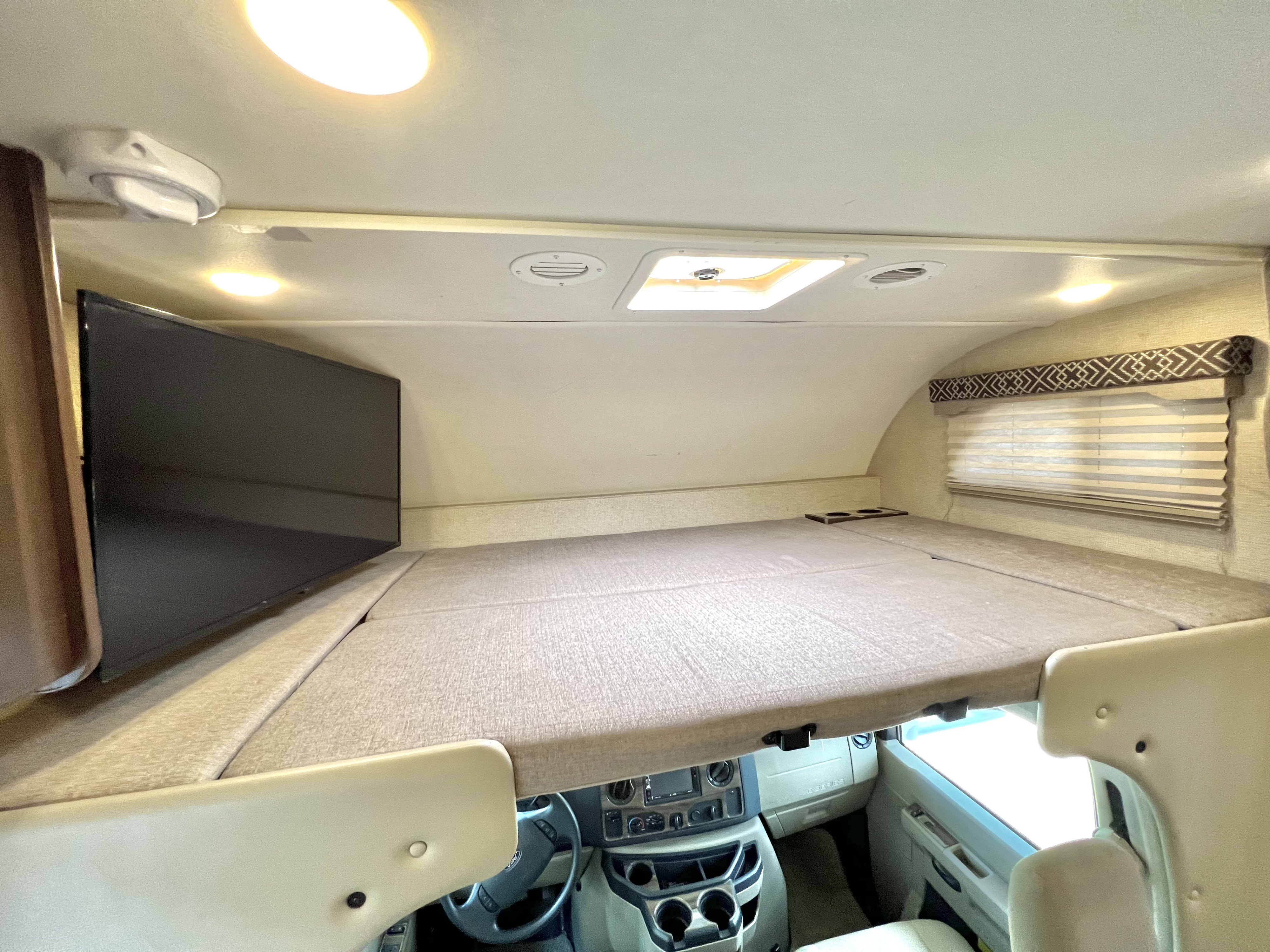 Inside an RV rental: a cozy cabover bunk with a mounted TV, window, and skylight, above the driving cab.. Thor Four Winds 2018