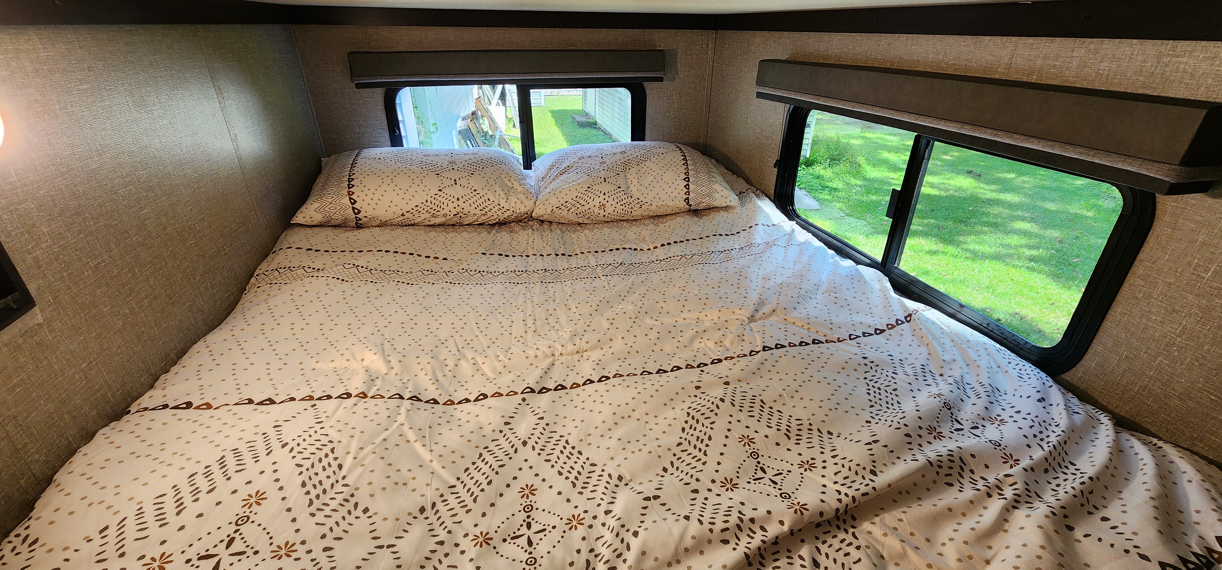 Cozy RV rental bedroom with a patterned bed, pillows, and bright windows showing green outdoors.. Grand Design Reflection 2021