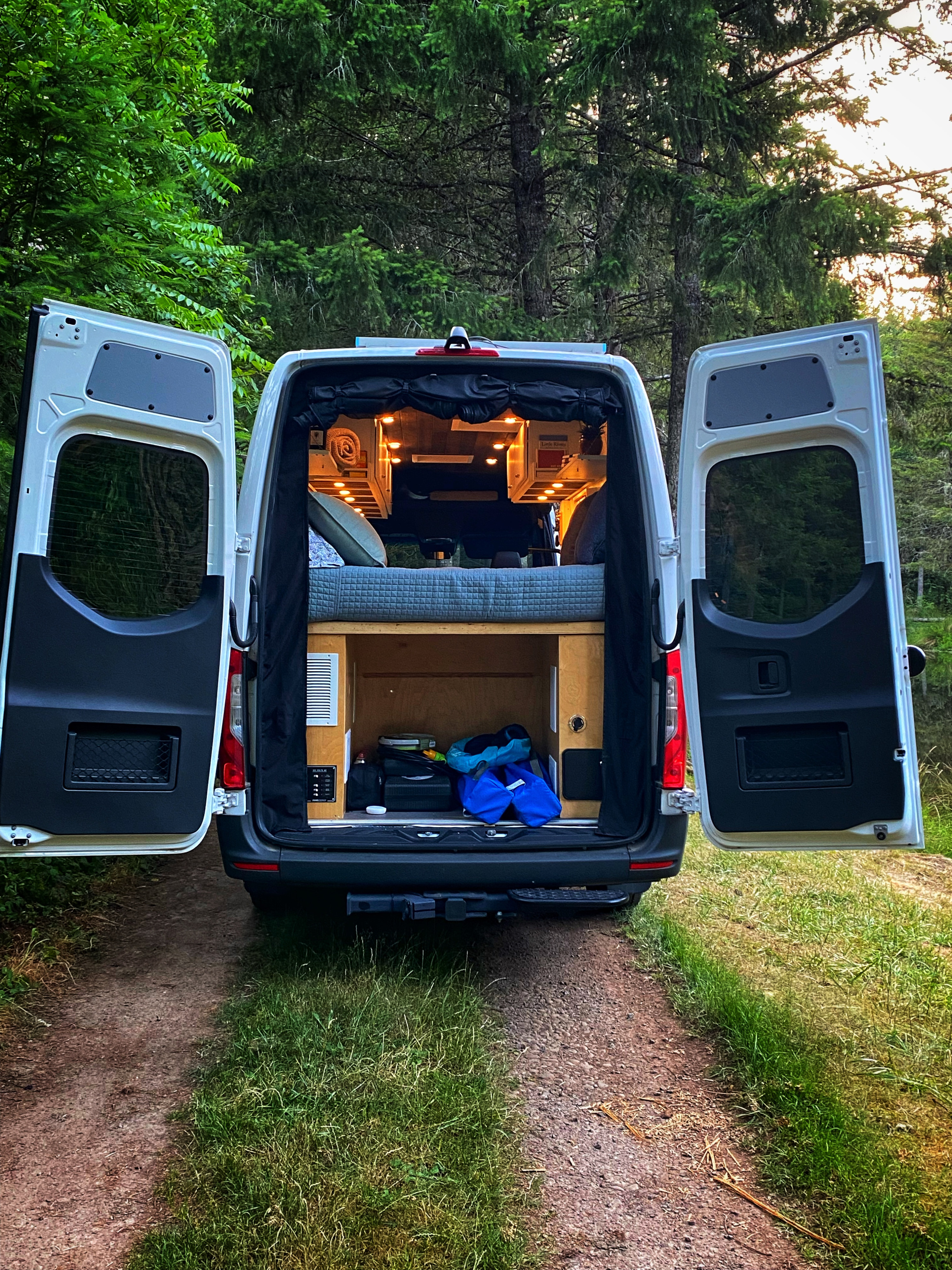 Rear doors of an RV rental open to reveal a comfortable bed and storage inside, set in a peaceful wooded area.. Freightliner Sprinter 2020