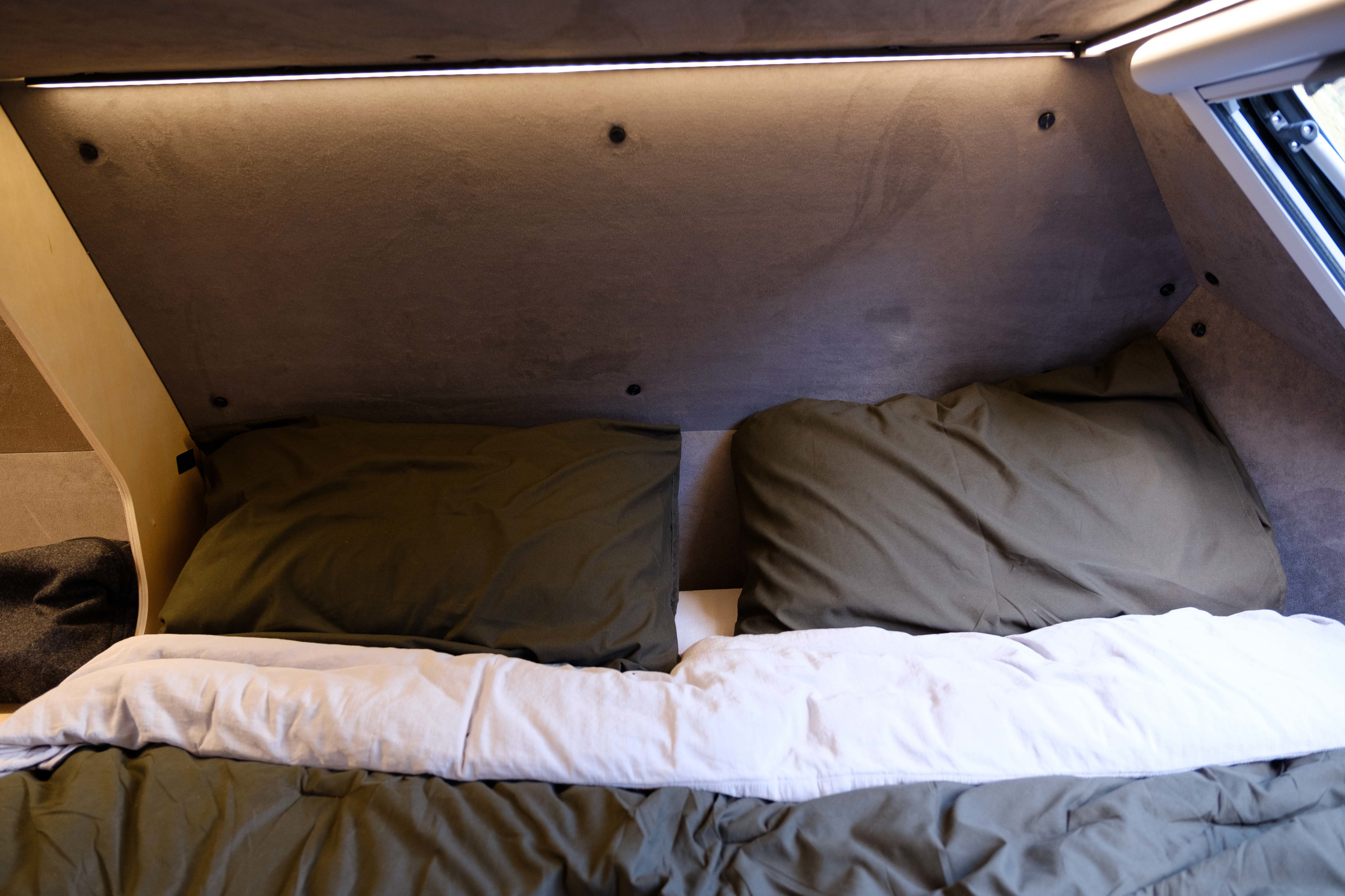 View inside an **rv rental**: a compact sleeping area with two olive green pillows, a white duvet, and overhead LED lighting.. Kimbo Camper Series 6 2023