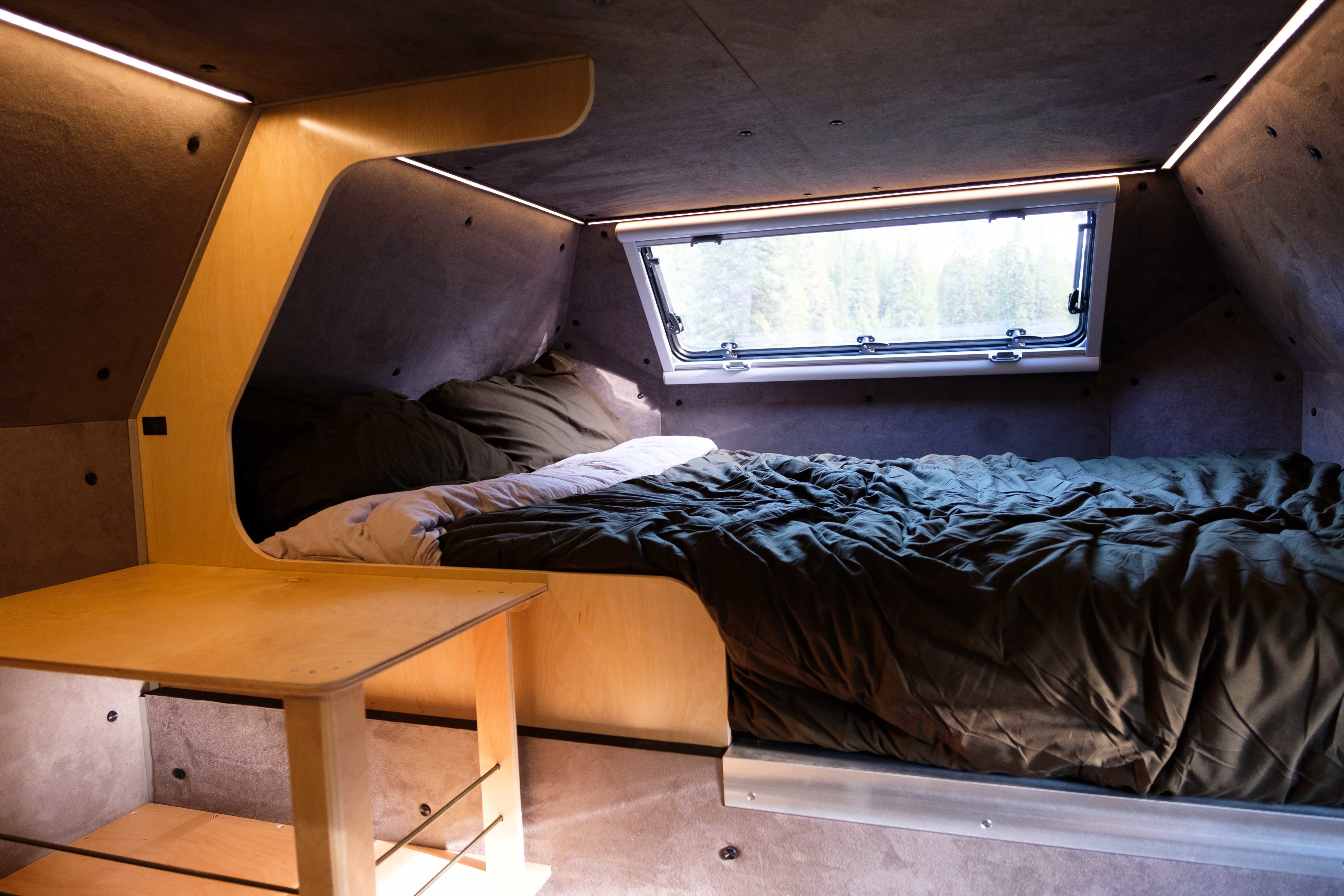 Experience comfort in this cozy RV rental interior, showcasing a compact bed, forest window view, and ambient lighting.. Kimbo Camper Series 6 2023