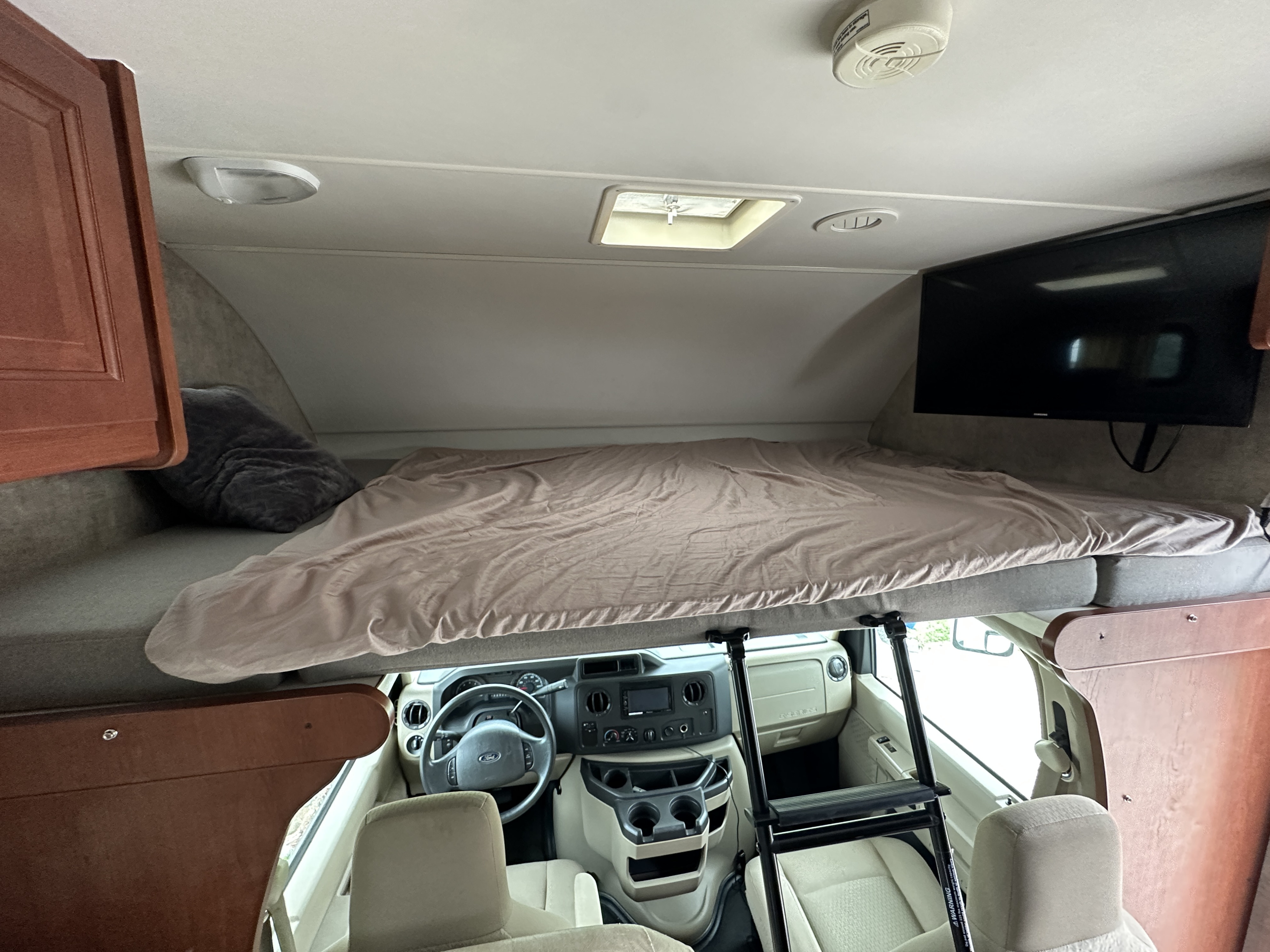 This RV rental interior showcases a cozy cab-over bed with a ladder, a TV, and the full driving area below.. Winnebago Minnie Winnie 2014