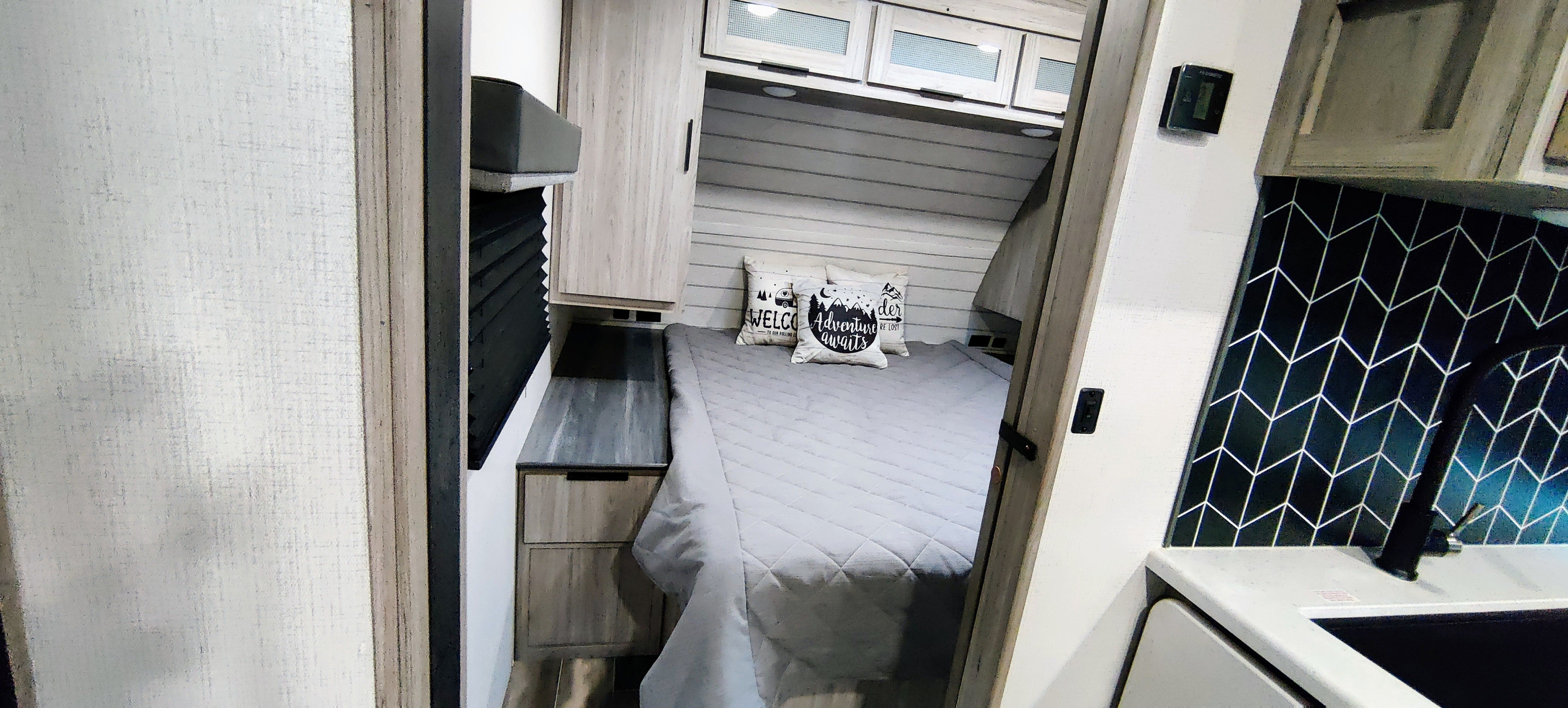 Explore this modern RV rental: a comfy bedroom with "Adventure Awaits" pillows, light wood cabinets, and a sleek kitchen backsplash.. Keystone RV Passport GT 2022