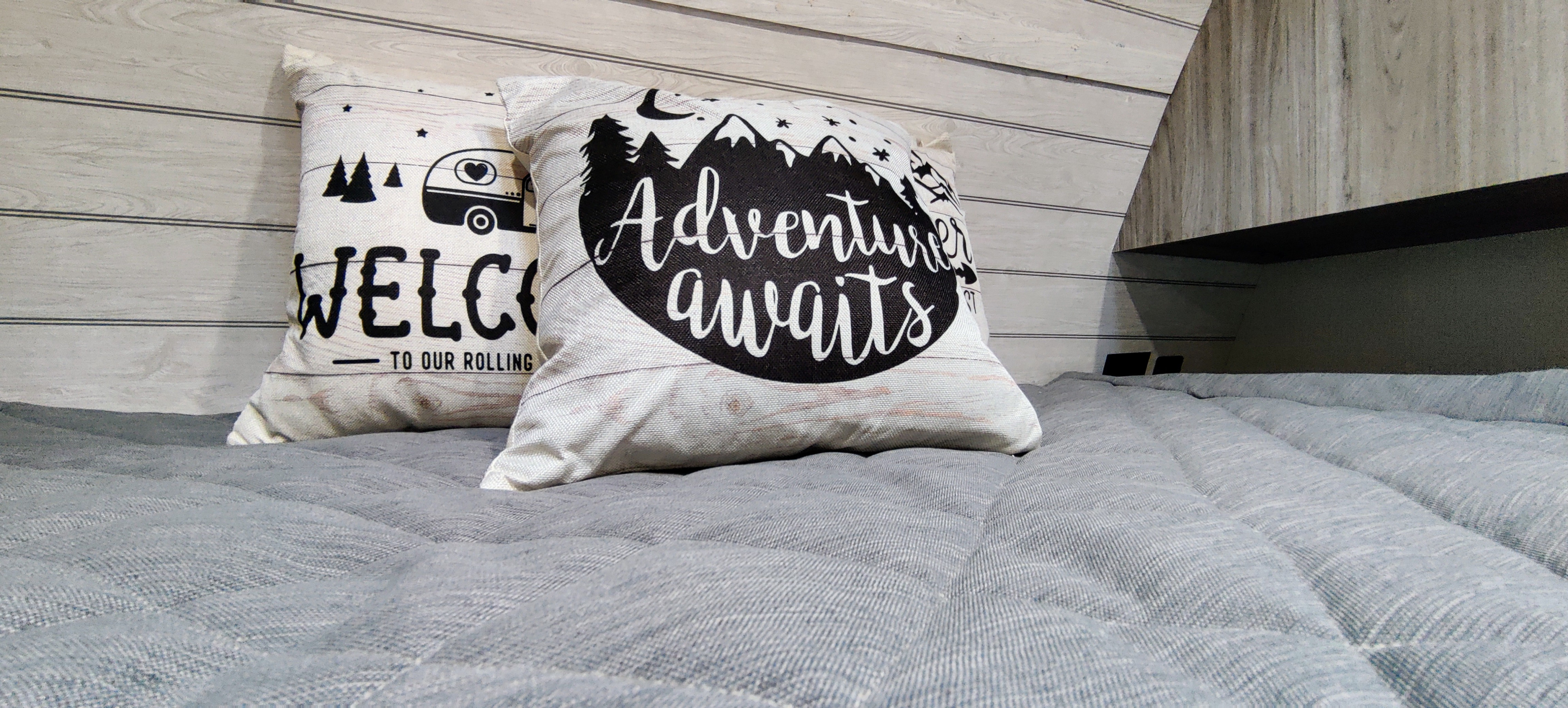 "Adventure awaits" on decorative pillows in a cozy RV rental bedroom setting with a soft gray comforter.. Keystone RV Passport GT 2022