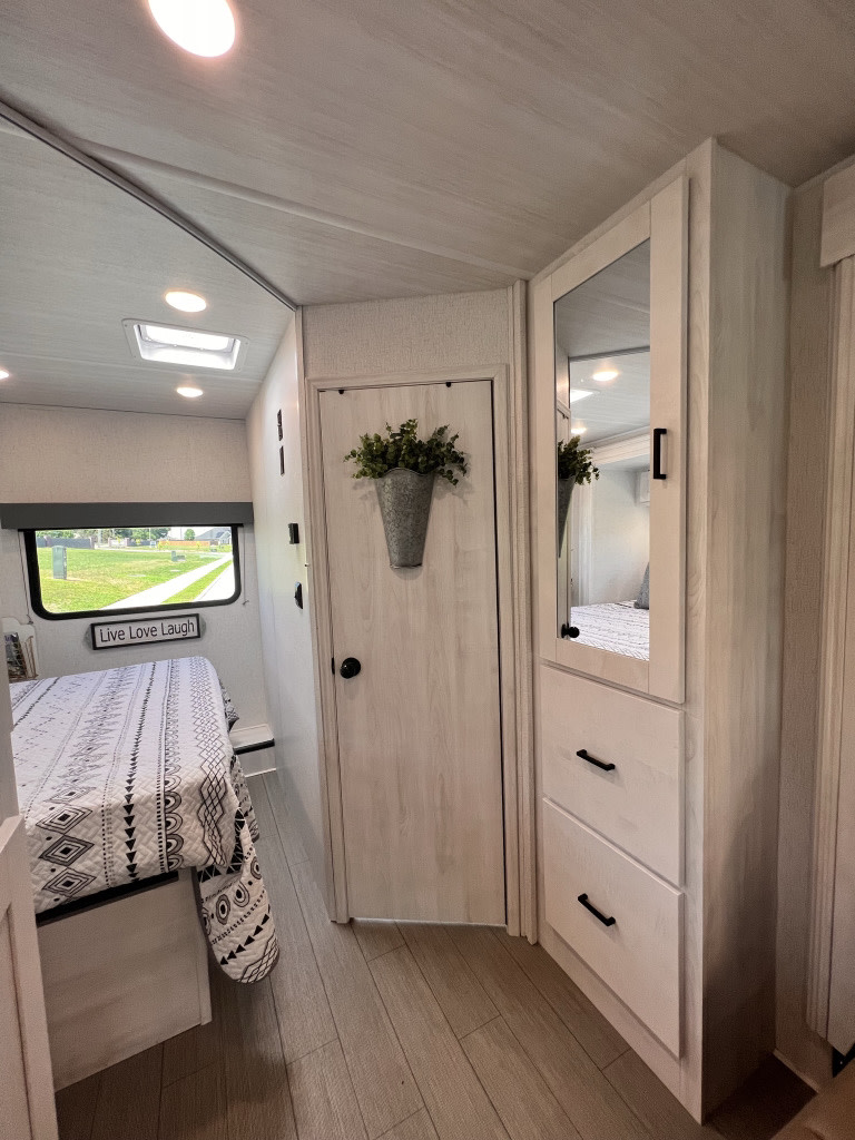 Bright RV rental interior: bedroom with patterned bedding, wood-look floors, modern storage, and mirrored cabinet.. East to West Entrada 2022
