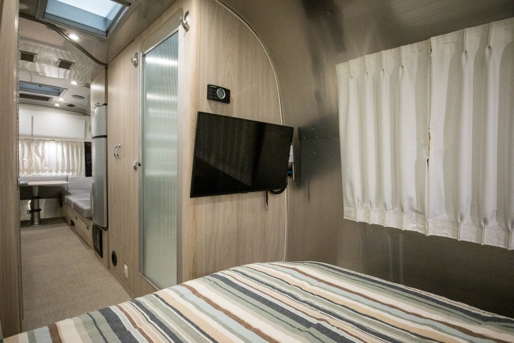 Cozy rv rental interior: A striped bed, mounted TV, sleek kitchen, and dining booth offer modern comfort on the road.. Airstream Globetrotter 2019