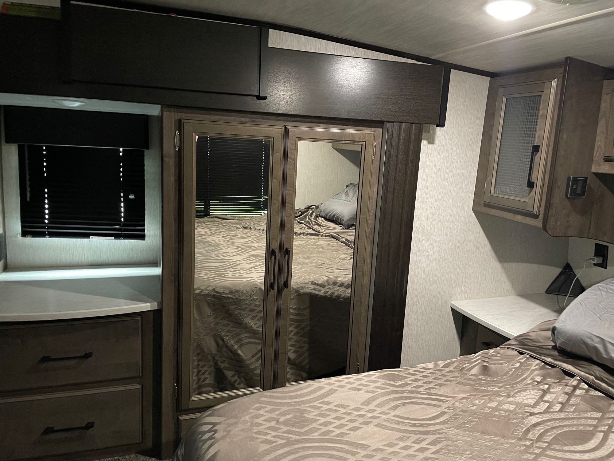 An RV rental bedroom featuring a patterned bedspread, mirrored closet, overhead cabinets, and a window with blinds.. Heartland Torque 2019
