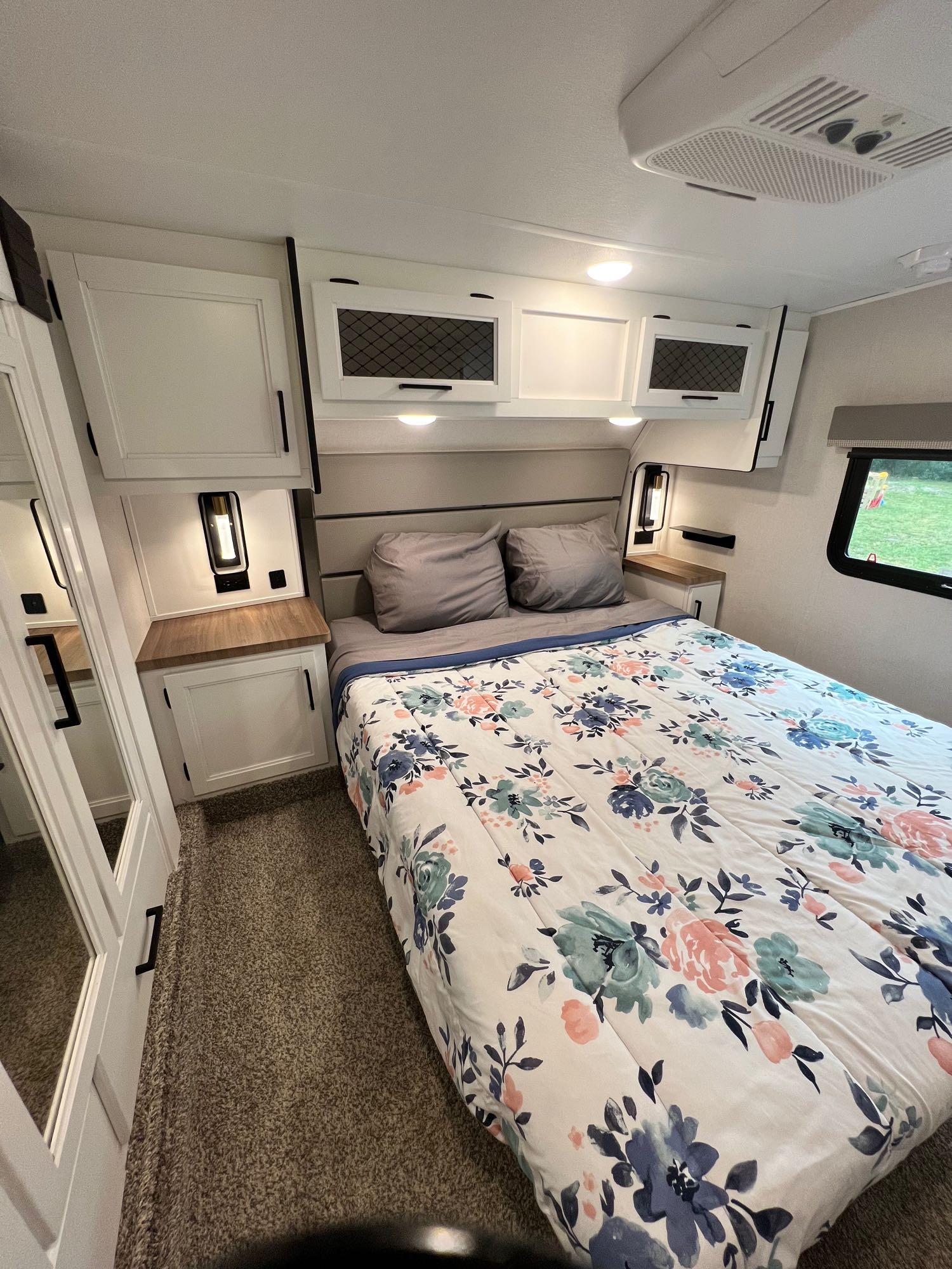 Cozy RV rental bedroom features a floral comforter, overhead storage, bedside lighting, and a window view, offering comfort.. Crusader Lite 2022