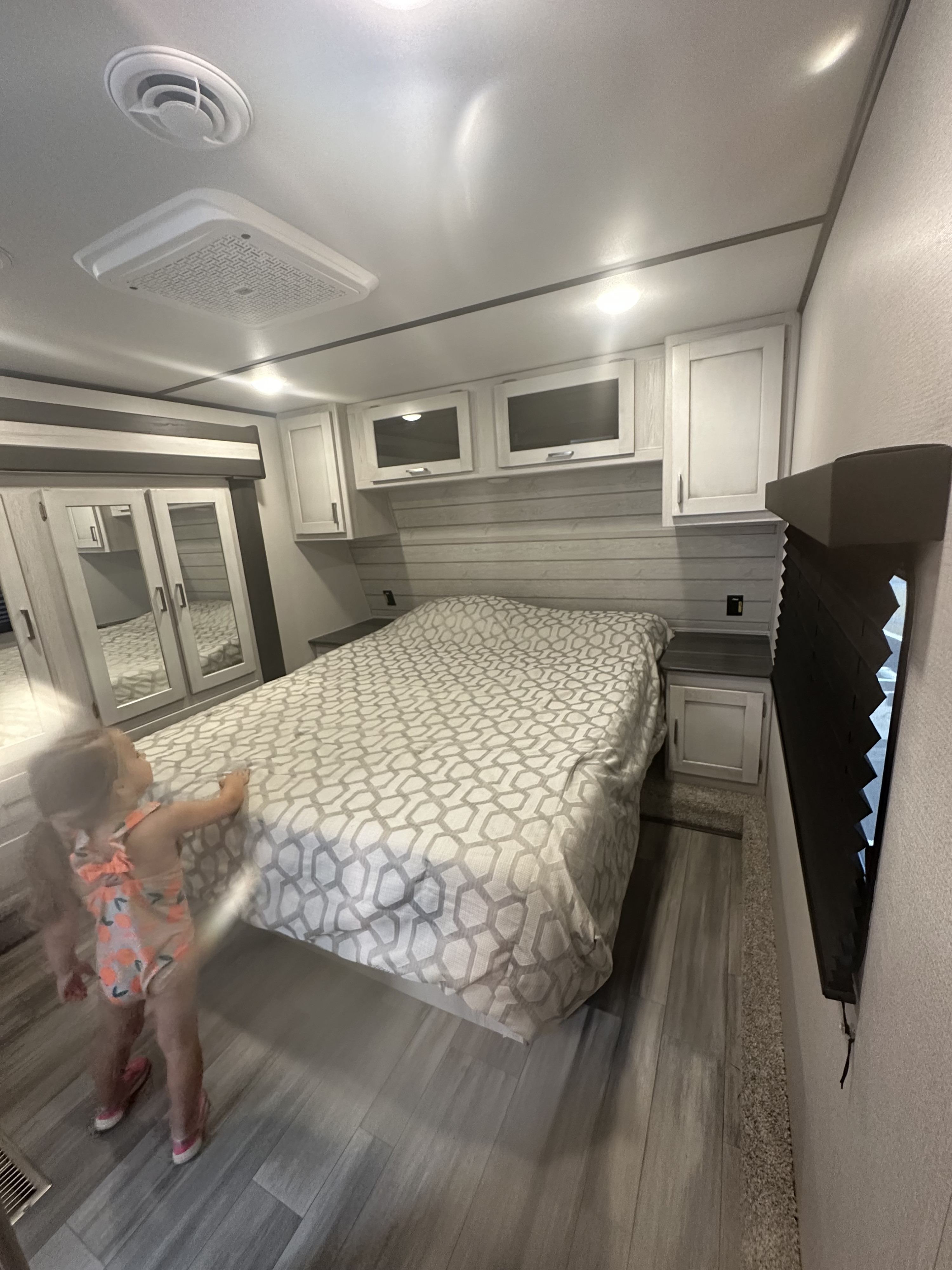 Child in a modern RV rental bedroom with patterned bed, overhead cabinets, mirrored closet, and wood-look floor.. Keystone RV Sprinter 2023