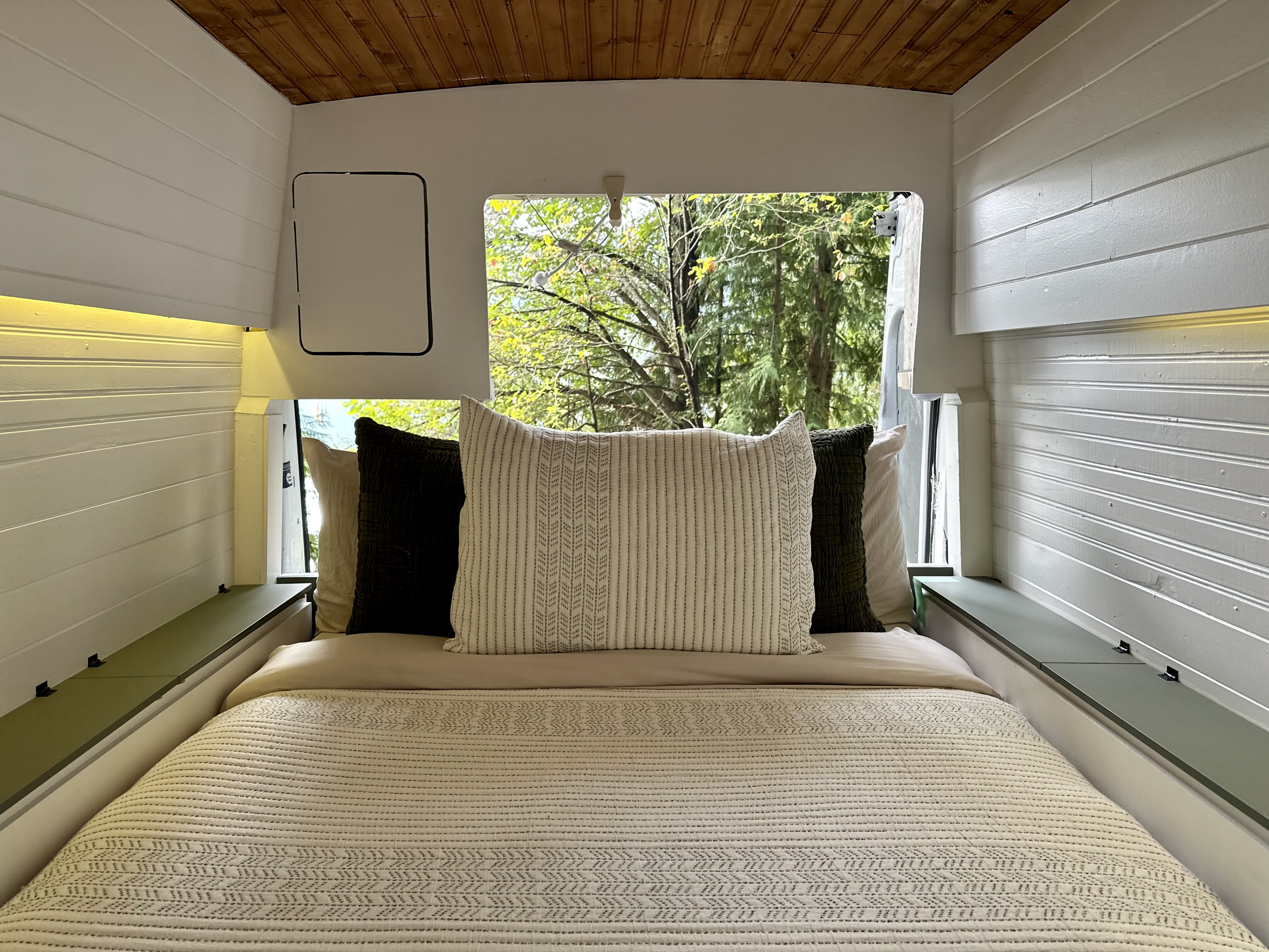 Relax in this RV rental bed area with cozy bedding, wood ceiling, and a large window showcasing nature. Integrated side storage adds function.. Ford Transit 2016