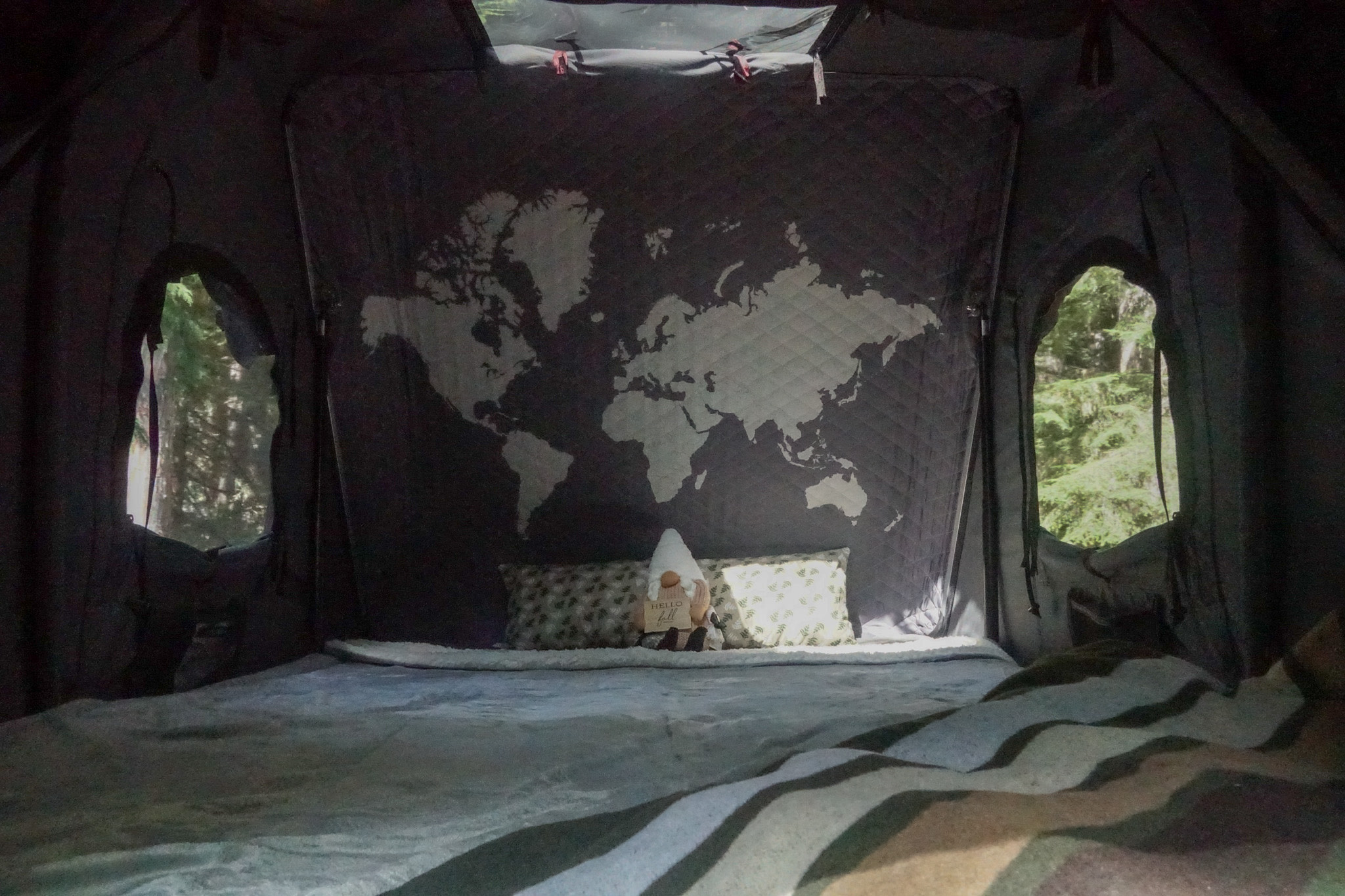 Cozy interior of an RV rental with a world map wall, a bed, and scenic forest views through the windows.. Subaru Outback with iKamper 2017