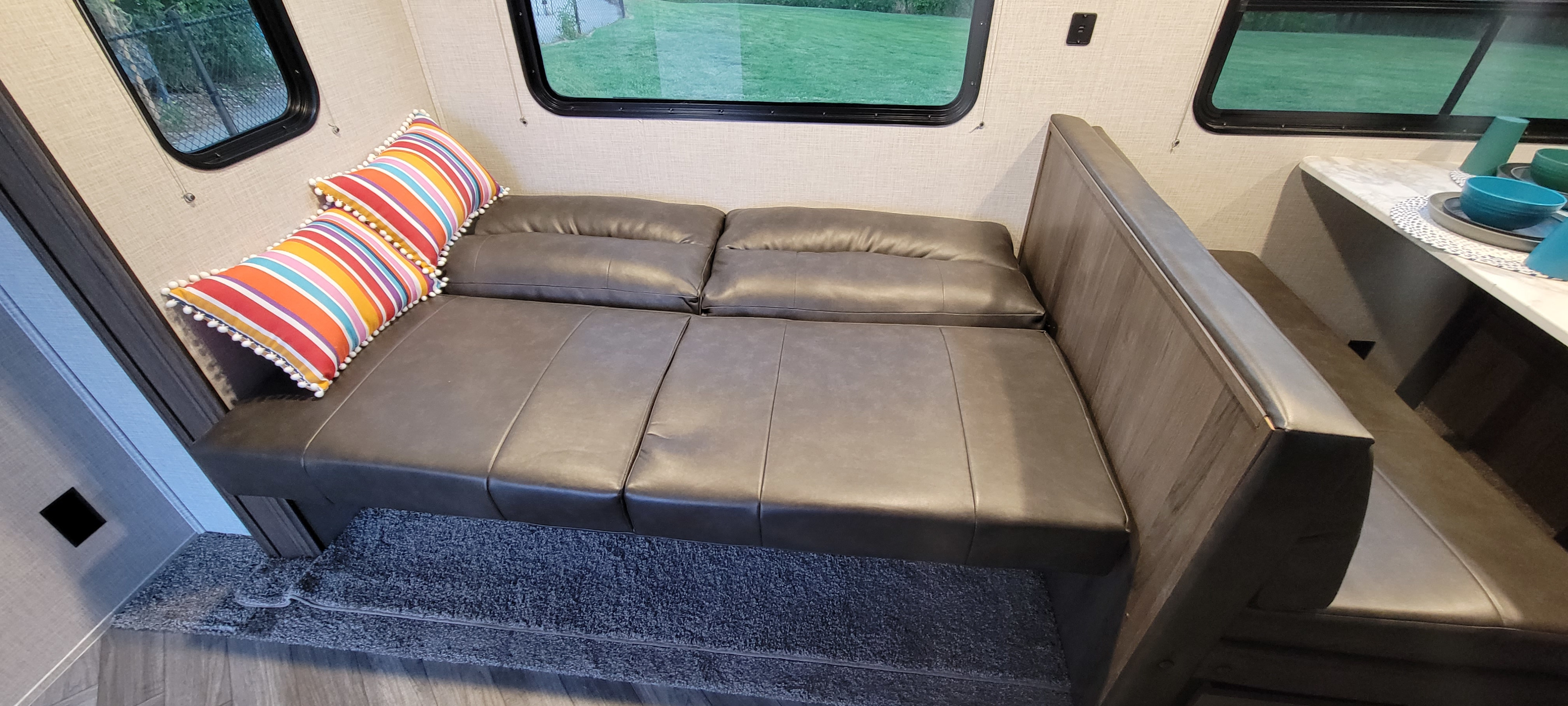 Spacious RV rental interior: gray faux leather convertible sofa/bed with colorful striped pillows, adjacent dinette, and large window view.. East to West Della Terra 2021