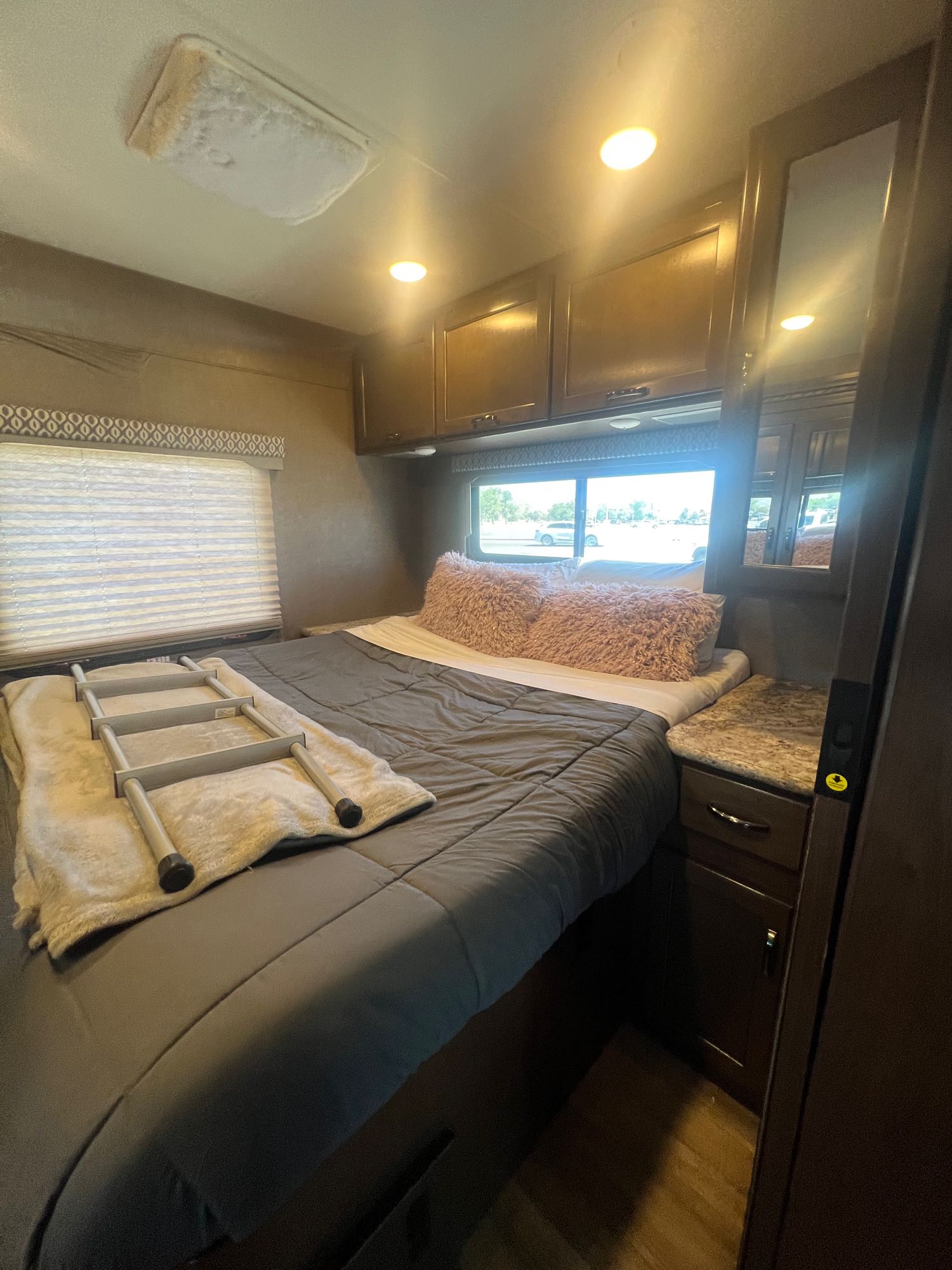 RV rental bedroom: a full bed with a ladder, overhead storage, bright lights, and window blinds revealing the outside.. Thor Freedom Elite 2017