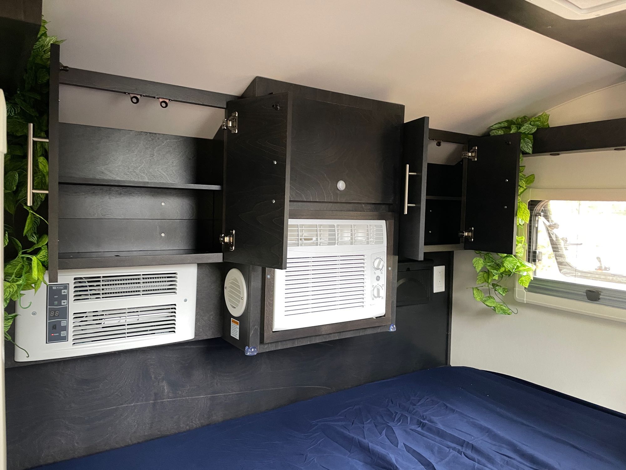 Modern RV rental bedroom featuring dark cabinetry, two integrated AC units, accent lighting, and green foliage above a blue bed.. nuCamp T@G 2023