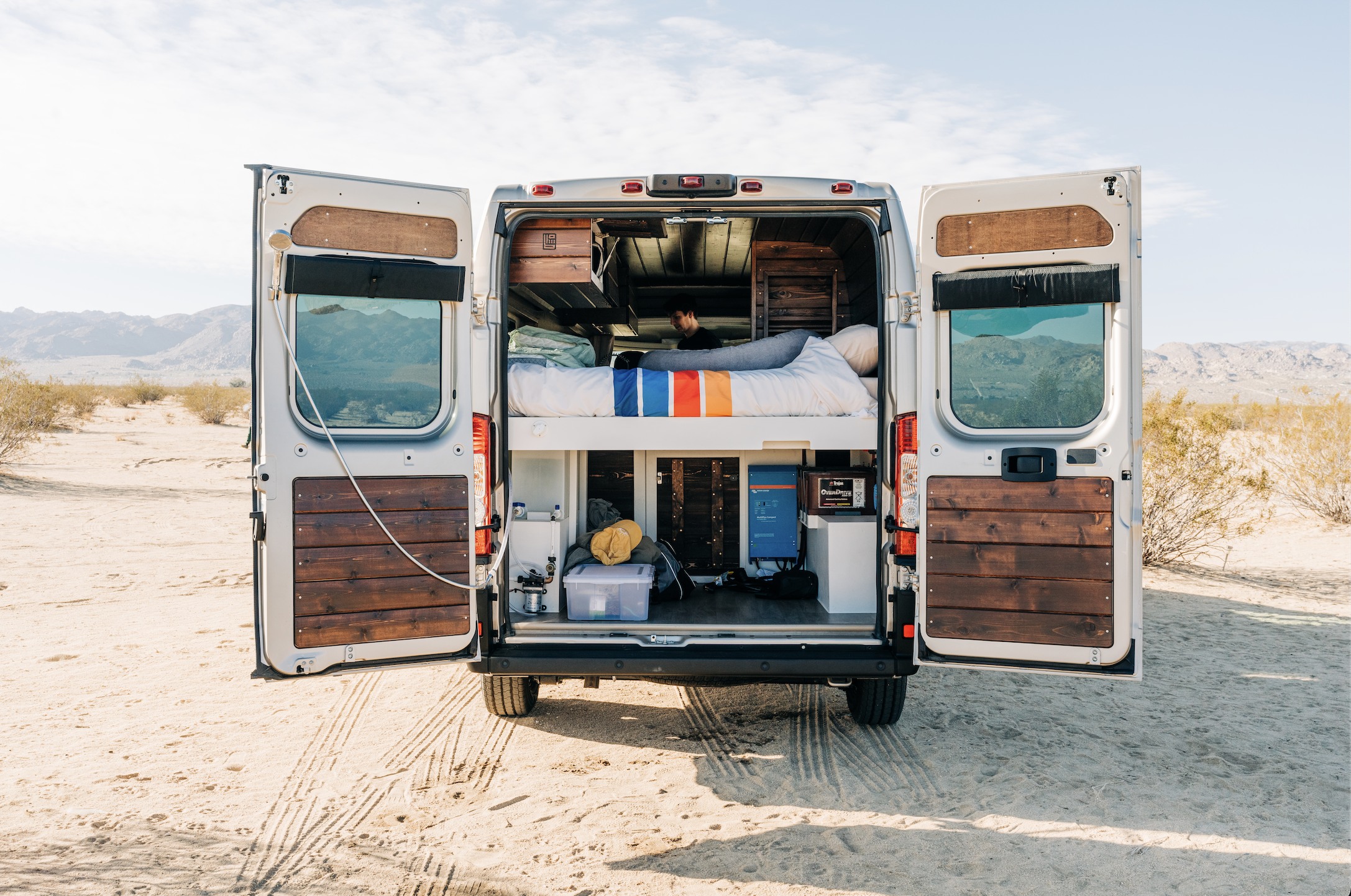 Rear view of an open rv rental camper van in the desert. Inside are a bed, storage, and utility setup. Mountains in background.. RAM Promaster 2021