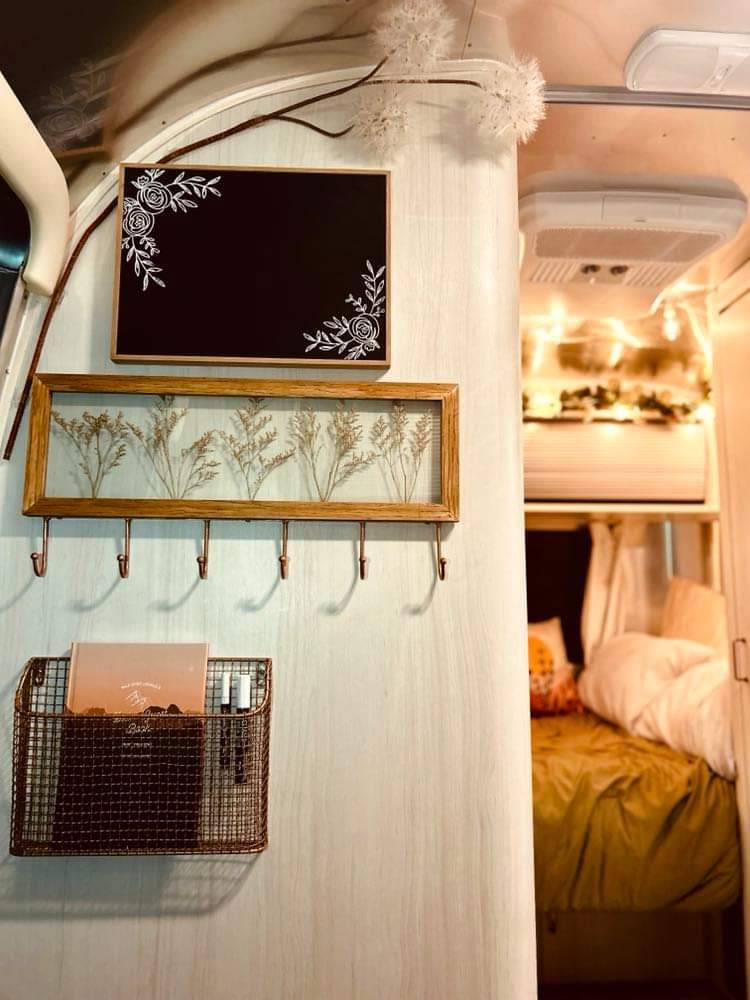 This RV rental interior features decorative signs, a pressed botanical frame with hooks, and a mesh basket, leading to the cozy bedroom.. Airstream Bambi 2021