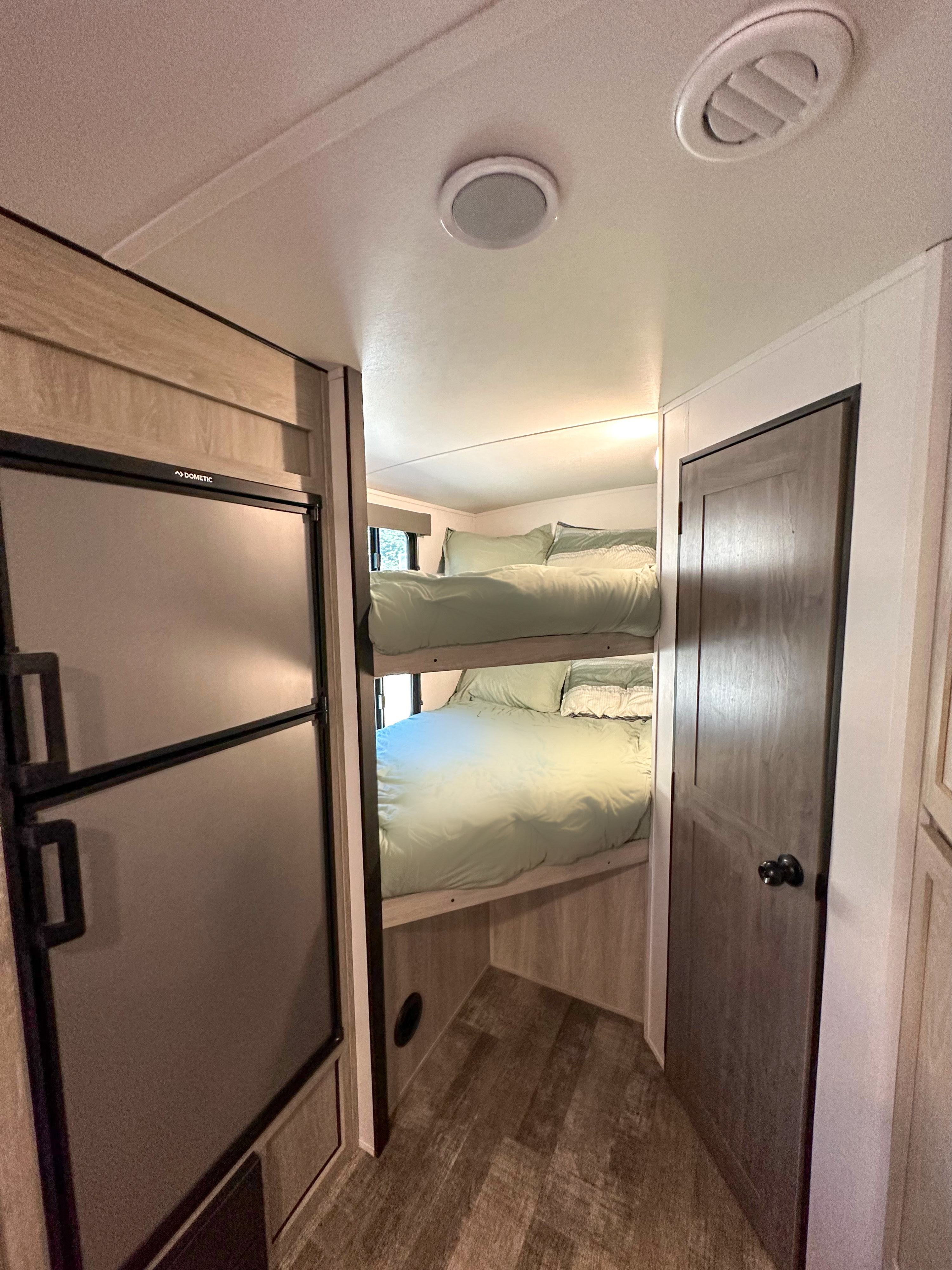 Inside an RV rental, showing bunk beds with light green bedding, a Dometic fridge, and a closed door, offering convenient amenities.. Riverside RV Xplorer 240BHx 2023