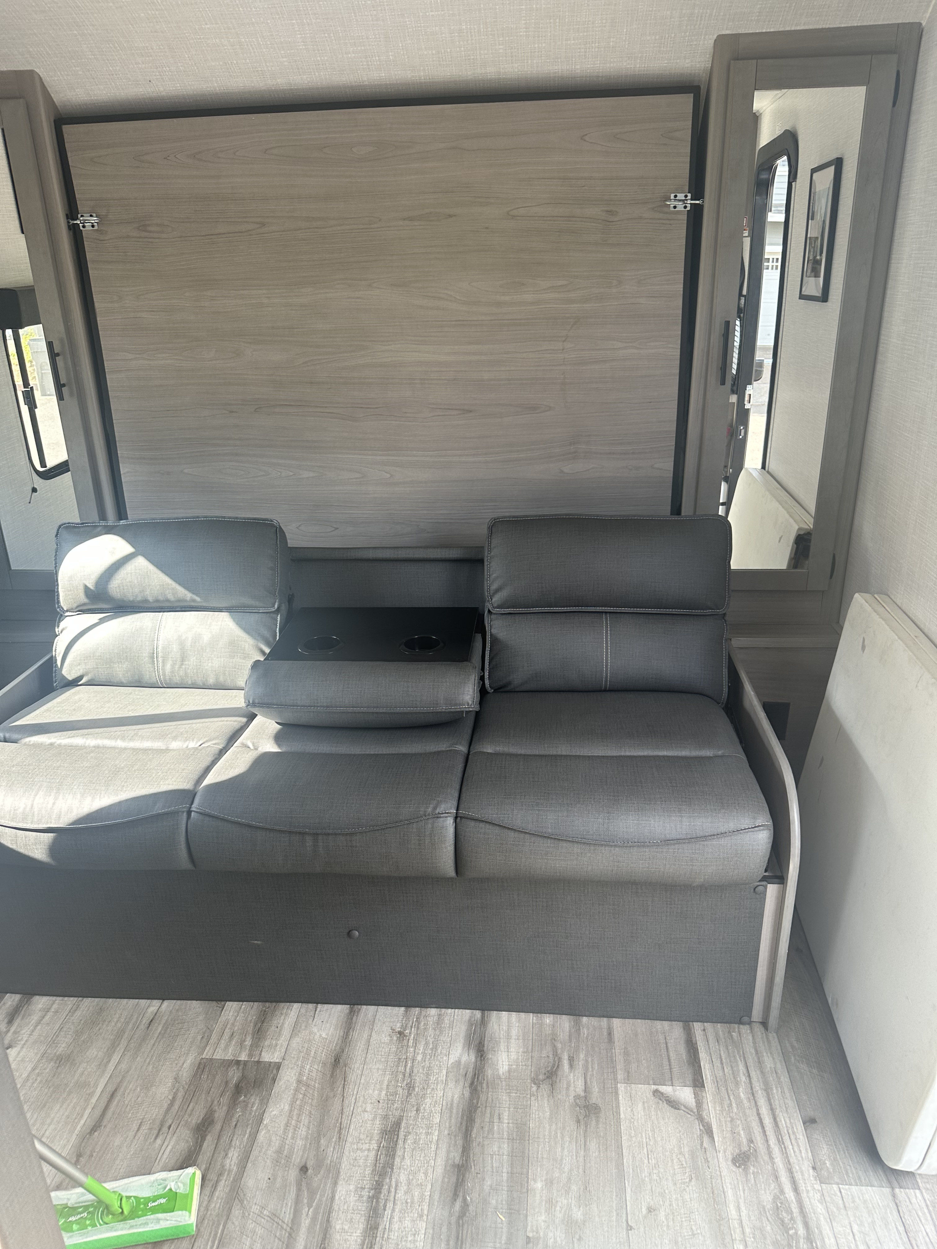 This RV rental interior features a grey sofa with built-in cup holders, facing a stylish wood-paneled wall, likely a stow-away bed.. Dutchmen Coleman 2022