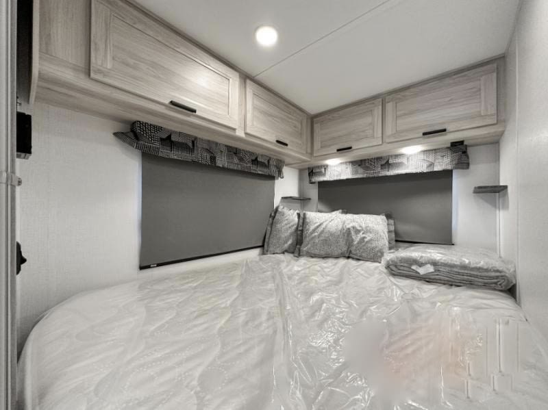 RV rental bedroom showing a protected mattress, pillows, light wood overhead cabinets, & grey window shades.. Forest River Sunseeker LE 2023