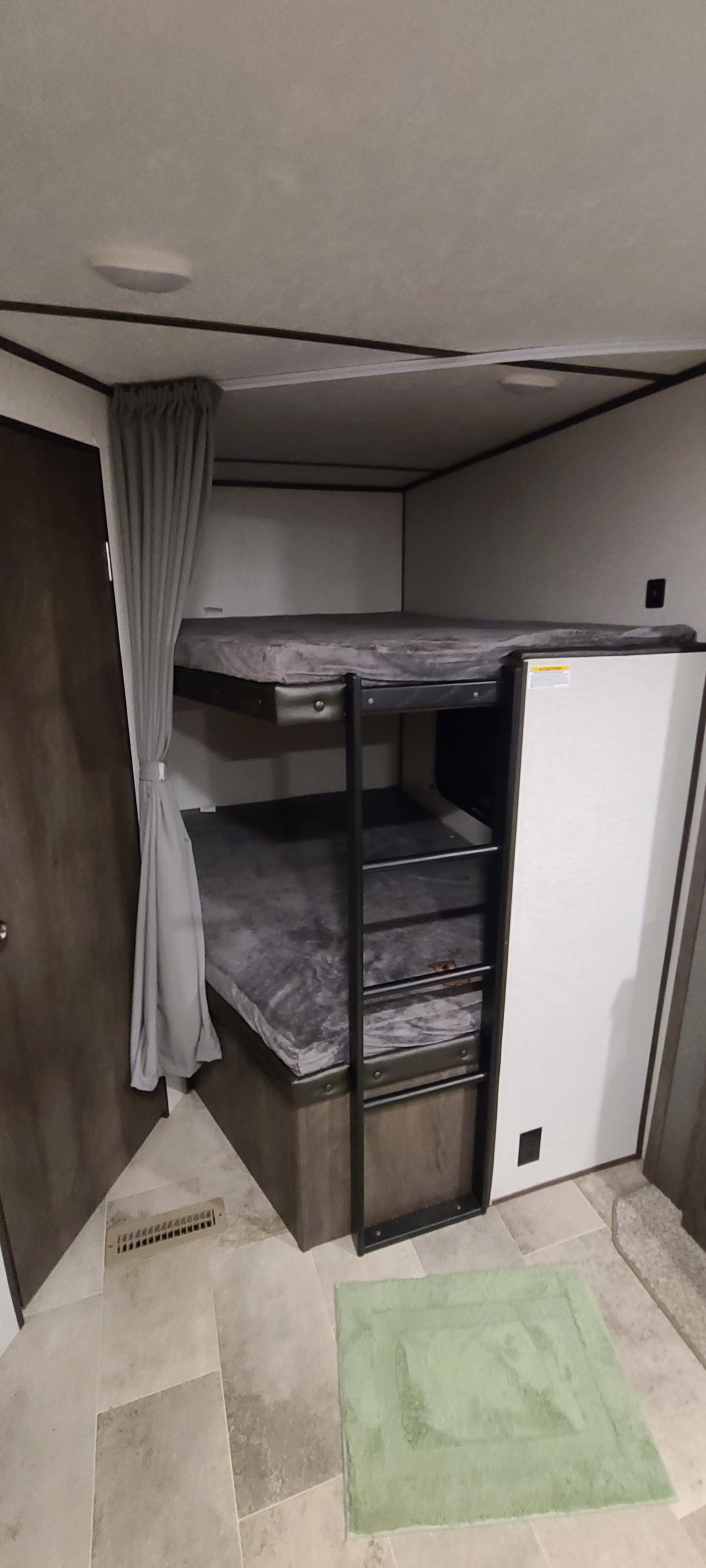 Cozy RV rental bunk beds, perfect for families. Features a ladder, privacy curtain, and light-toned flooring with a green mat.. Coleman Lantern 2021