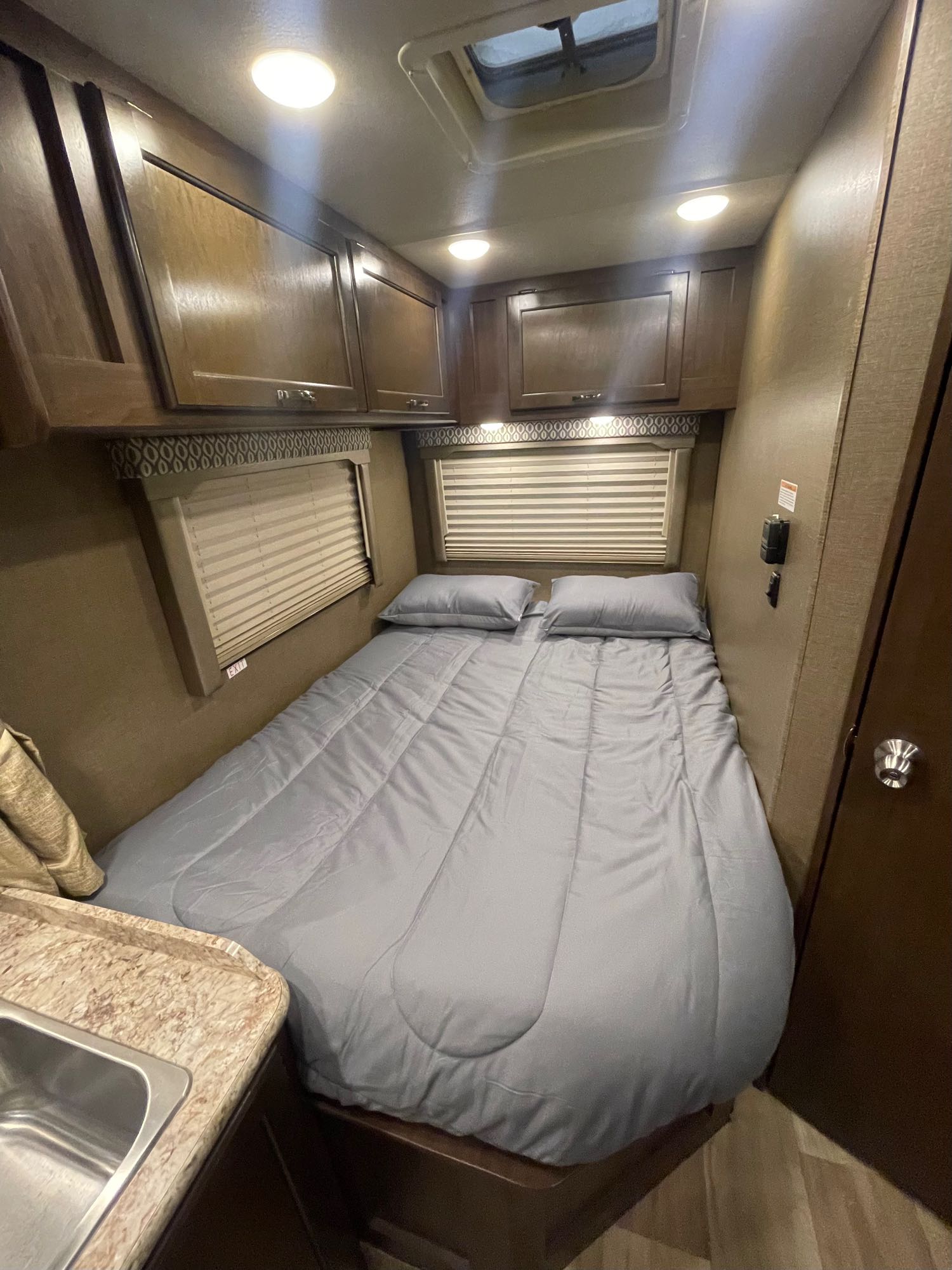 Cozy RV rental bedroom featuring a comfortable bed with gray bedding, overhead cabinets, and window blinds.. Thor Freedom Elite 2018