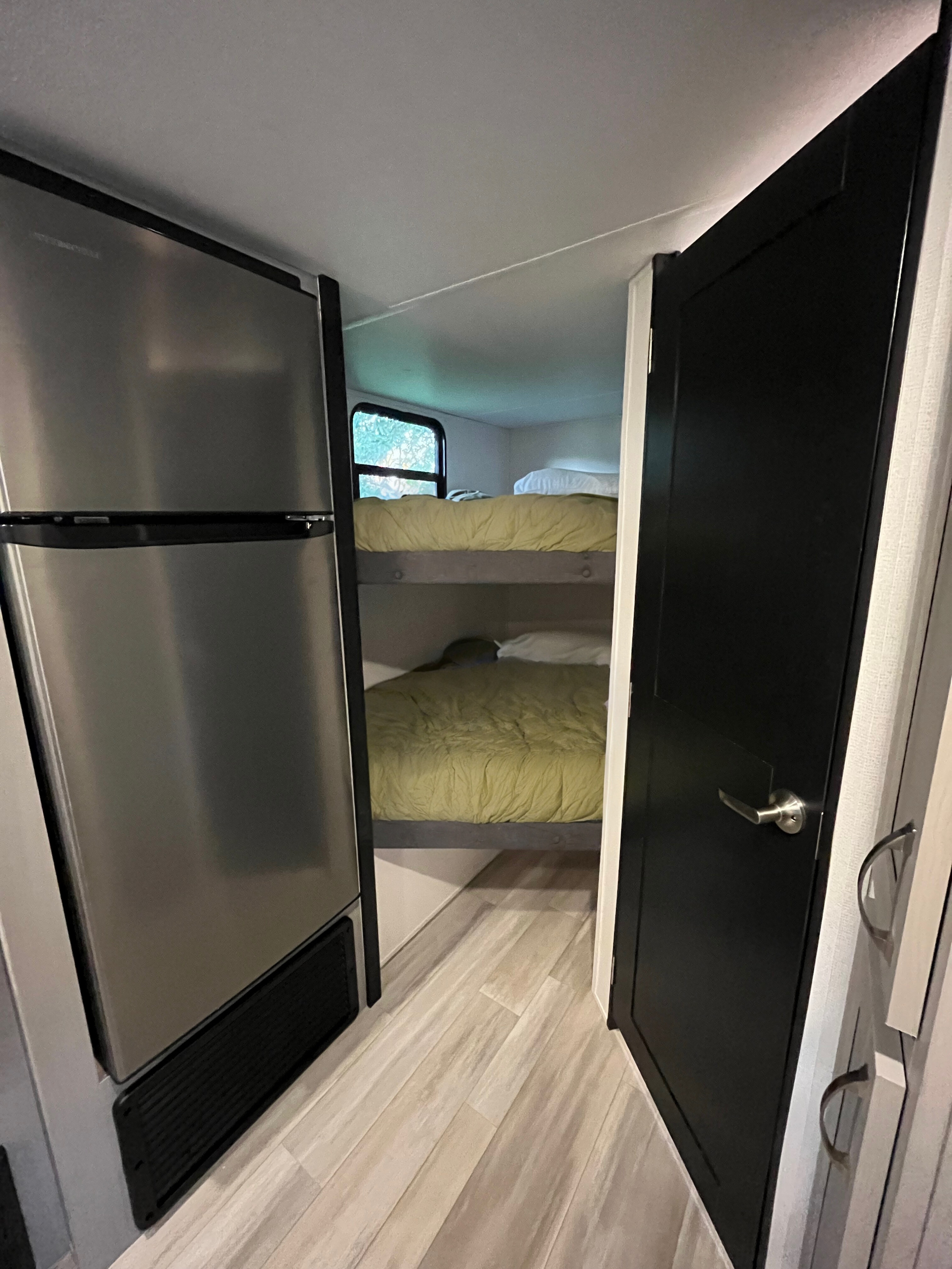 RV rental interior: Bunk beds with green bedding, a stainless steel refrigerator, and a black door on light wood flooring.. Forest River Tracer 2022