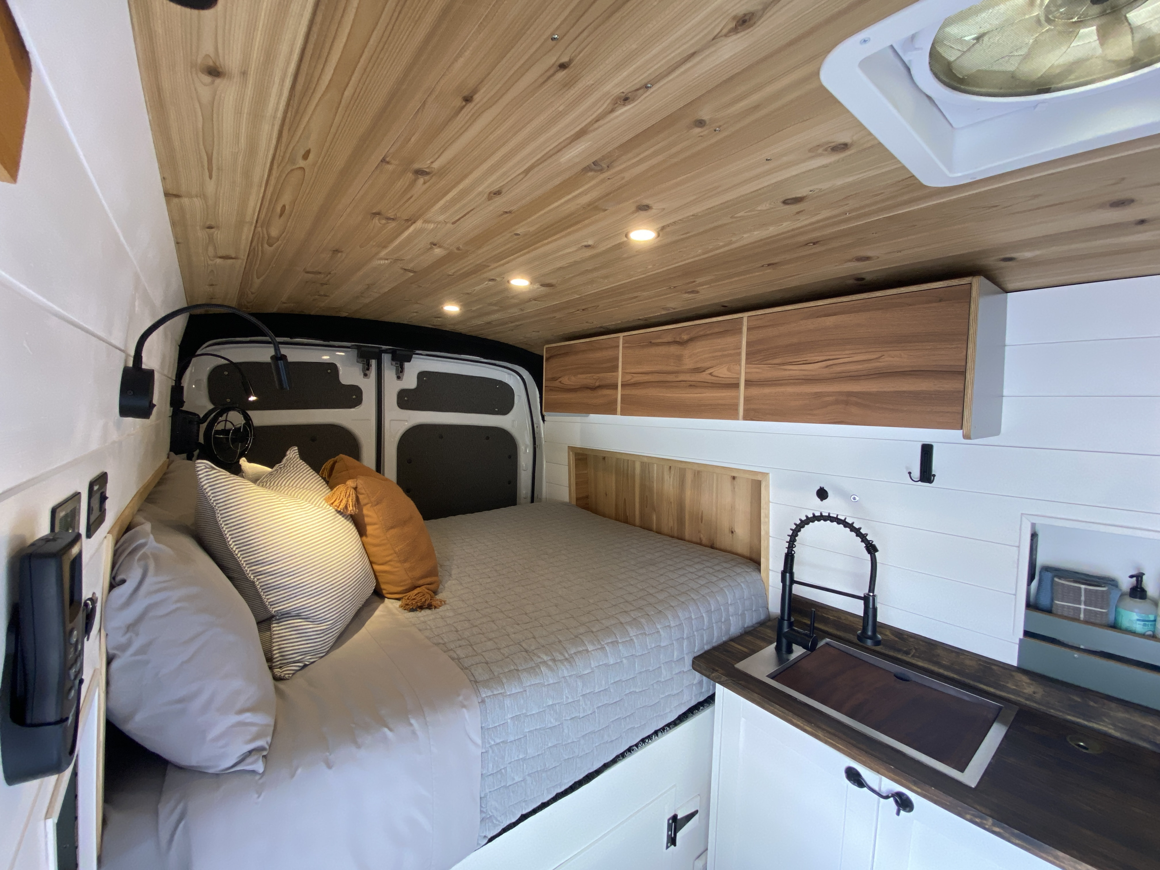 Modern RV rental interior showcasing a cozy bed with pillows, wooden ceiling, compact kitchen sink, and overhead cabinets.. Ford Transit custom 2019