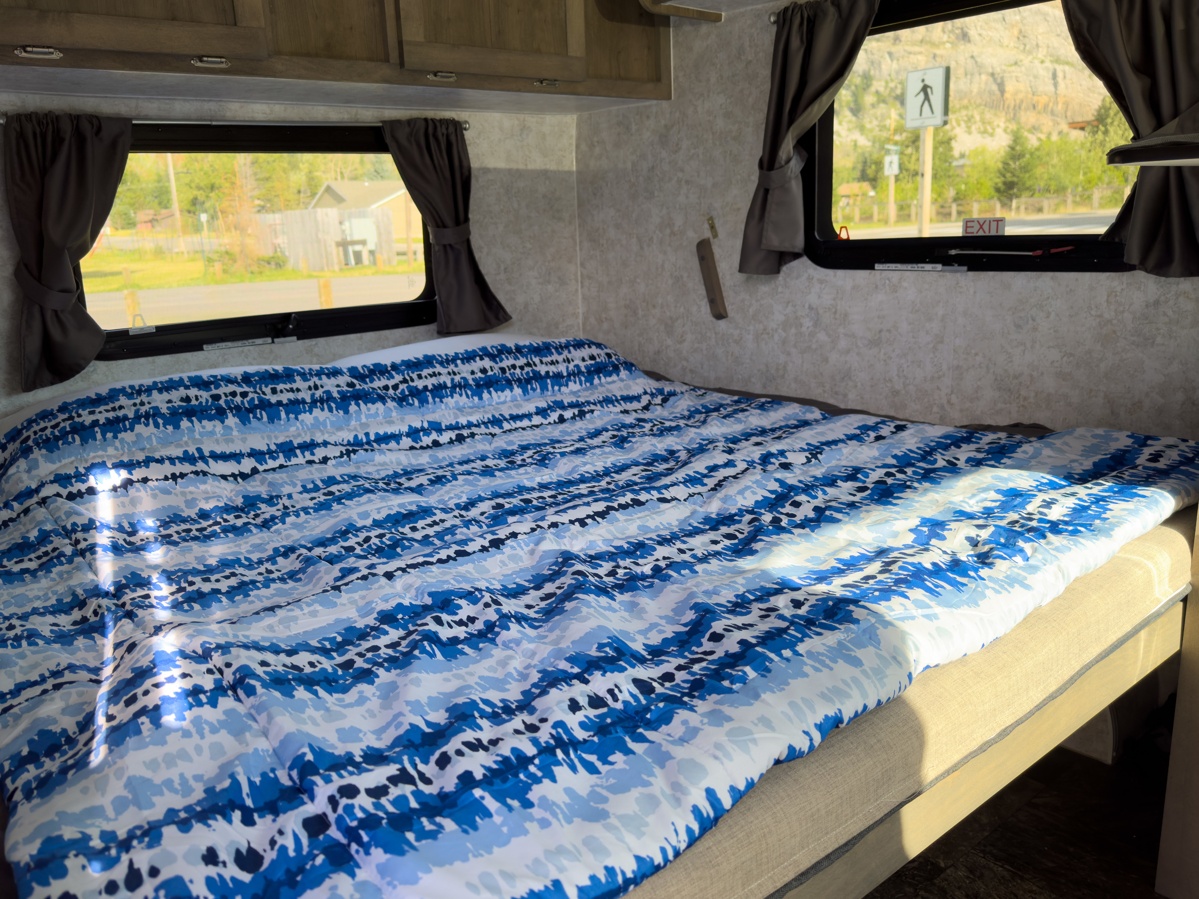 An RV rental bedroom with a blue patterned comforter on the bed, dark curtains, and a scenic view outside through the windows.. Prolite 12V 2023