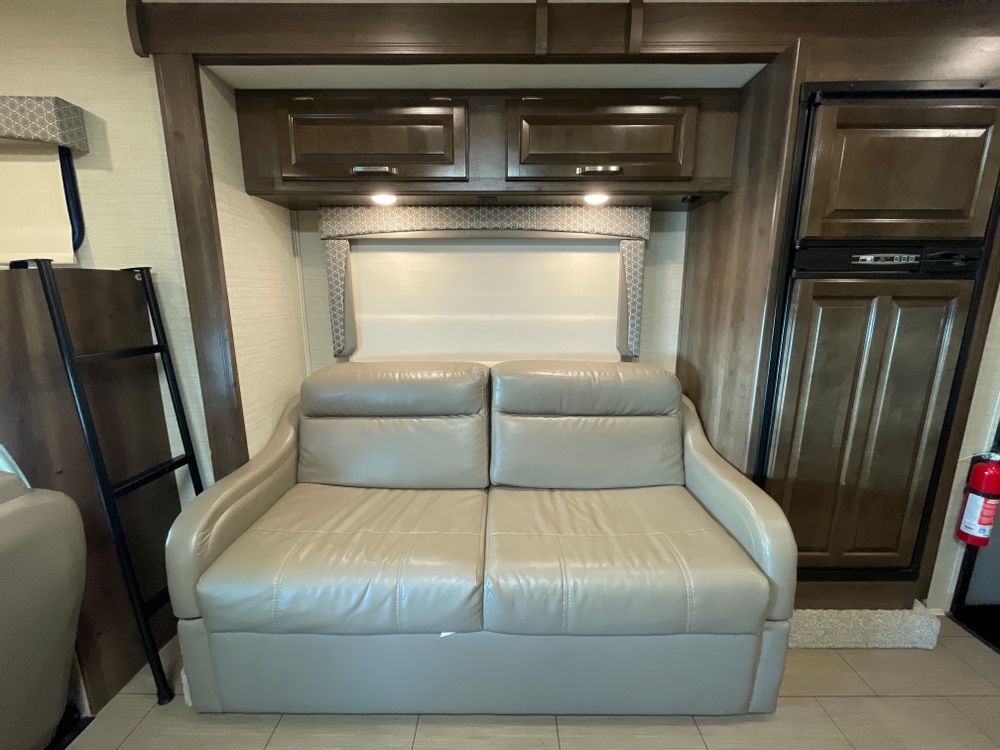 This RV rental interior shows a light-colored sofa below wood cabinets with lights. A ladder suggests bunks on the left, fridge on the right.. Jayco Greyhawk 2018