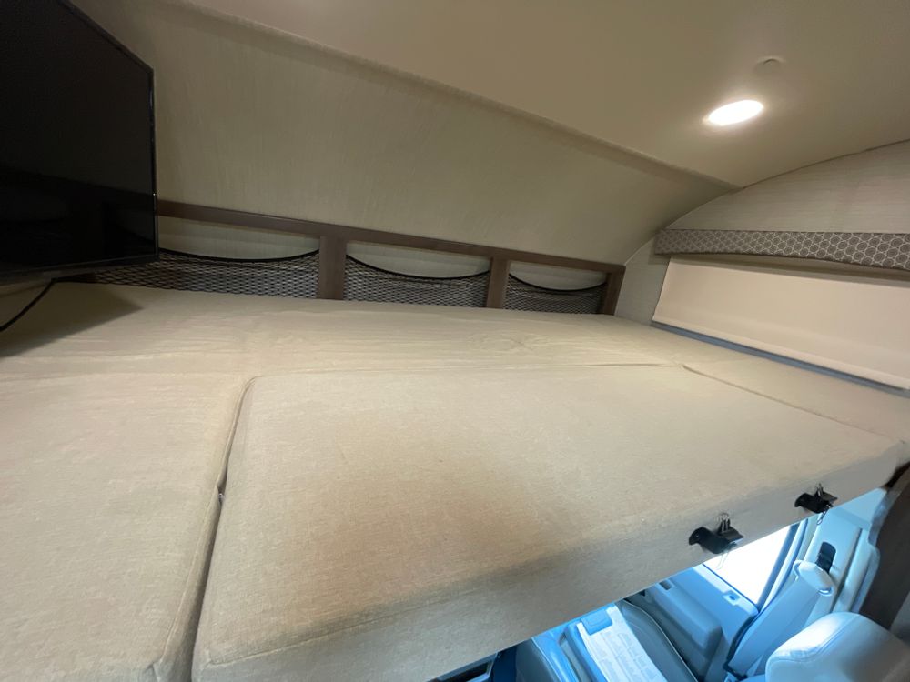 An RV rental's over-cab bunk, featuring a spacious bed, TV, storage nets, and window. Ideal sleeping area!. Jayco Greyhawk 2018