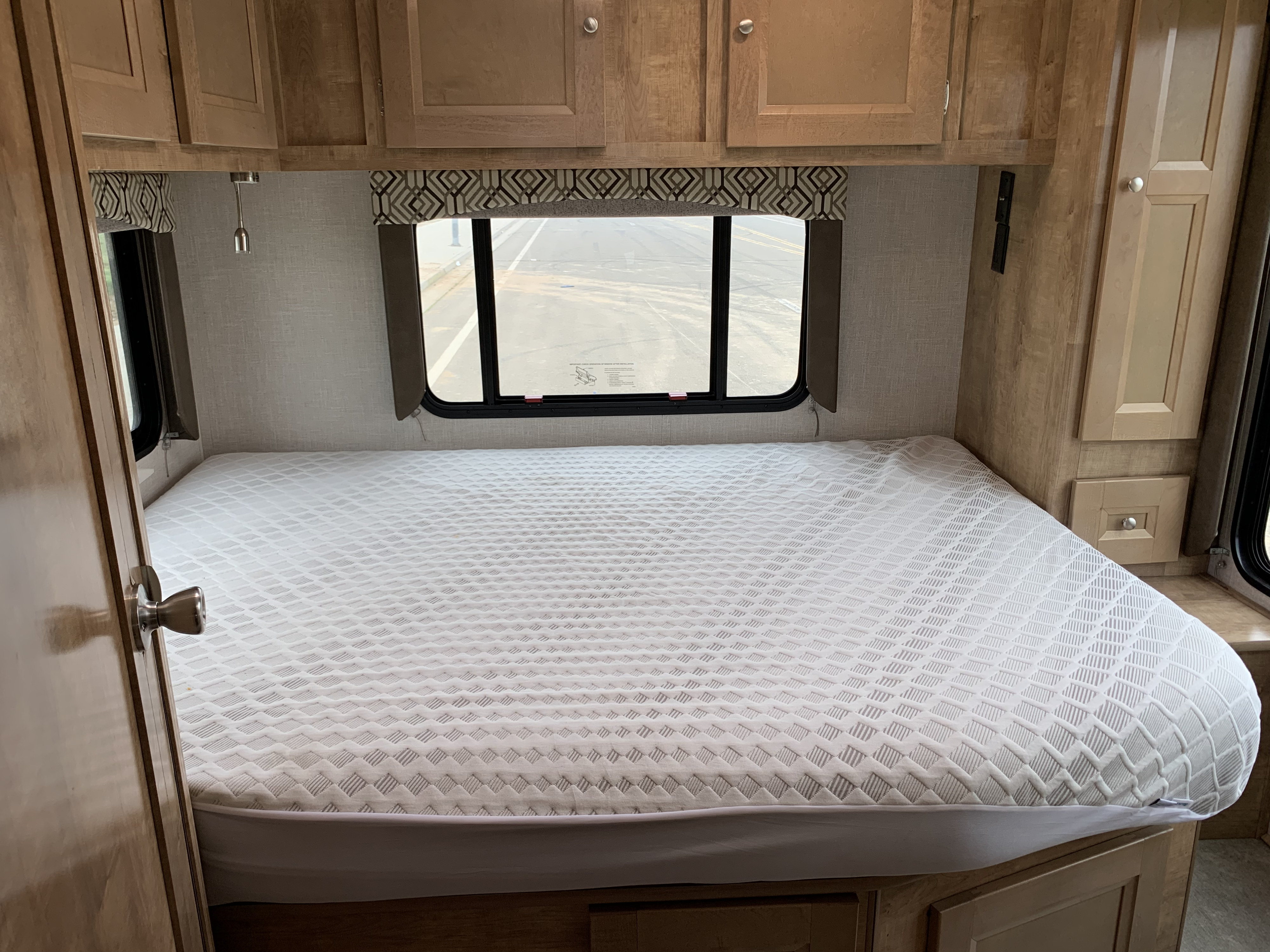 Clean, compact RV rental bedroom with white mattress, wooden cabinets, and a window offering a view outside.. Forest River Coachmen Leprechaun 2020