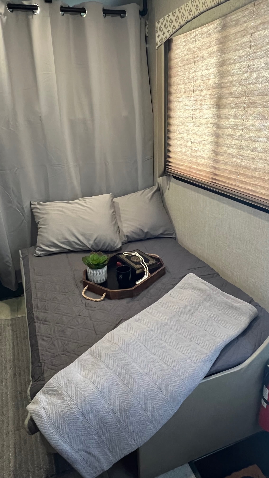 Inside a cozy RV rental: a bed with grey bedding, decorative tray, curtains, and a window with blinds for your journey.. Coachmen Freelander 2020