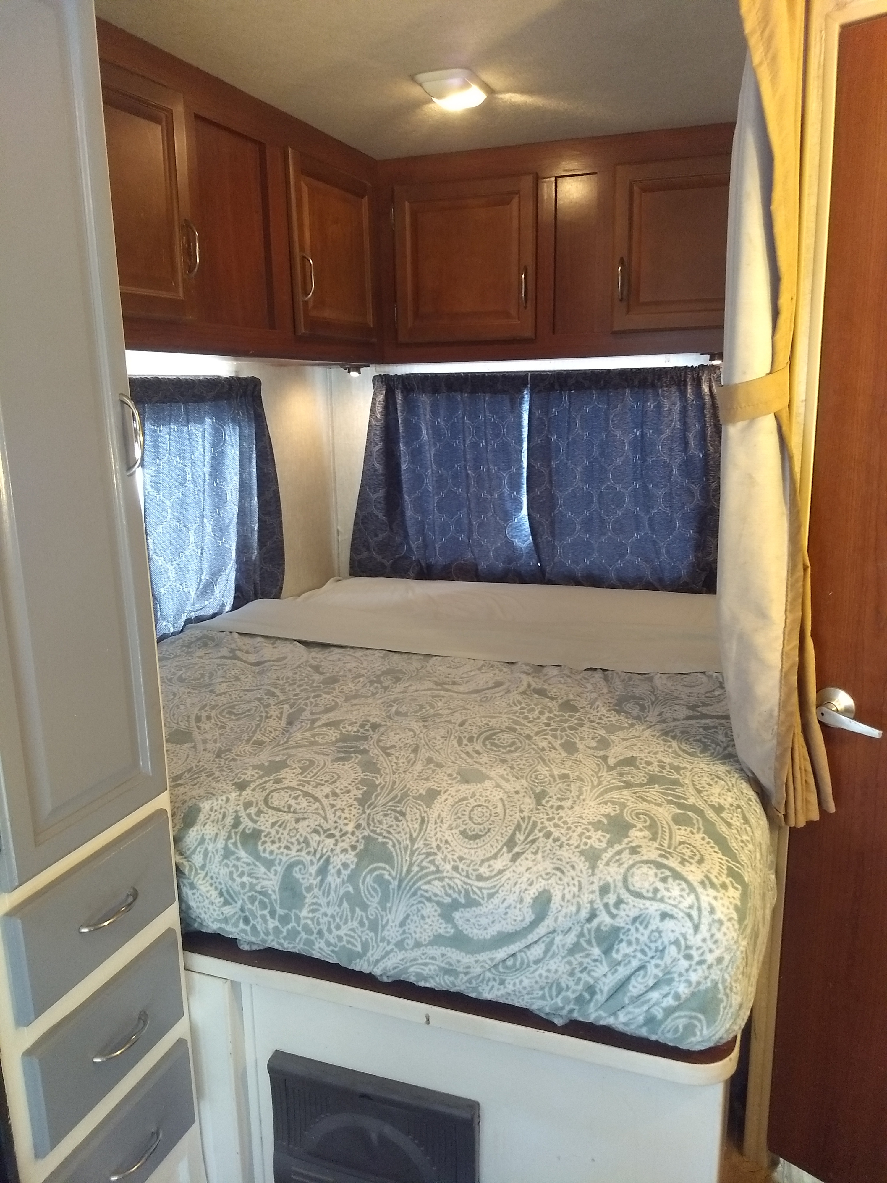 Cozy RV rental bedroom featuring a patterned bed, dark wood cabinets, and blue window curtains for a comfortable stay.. Fleetwood Jamboree 2009