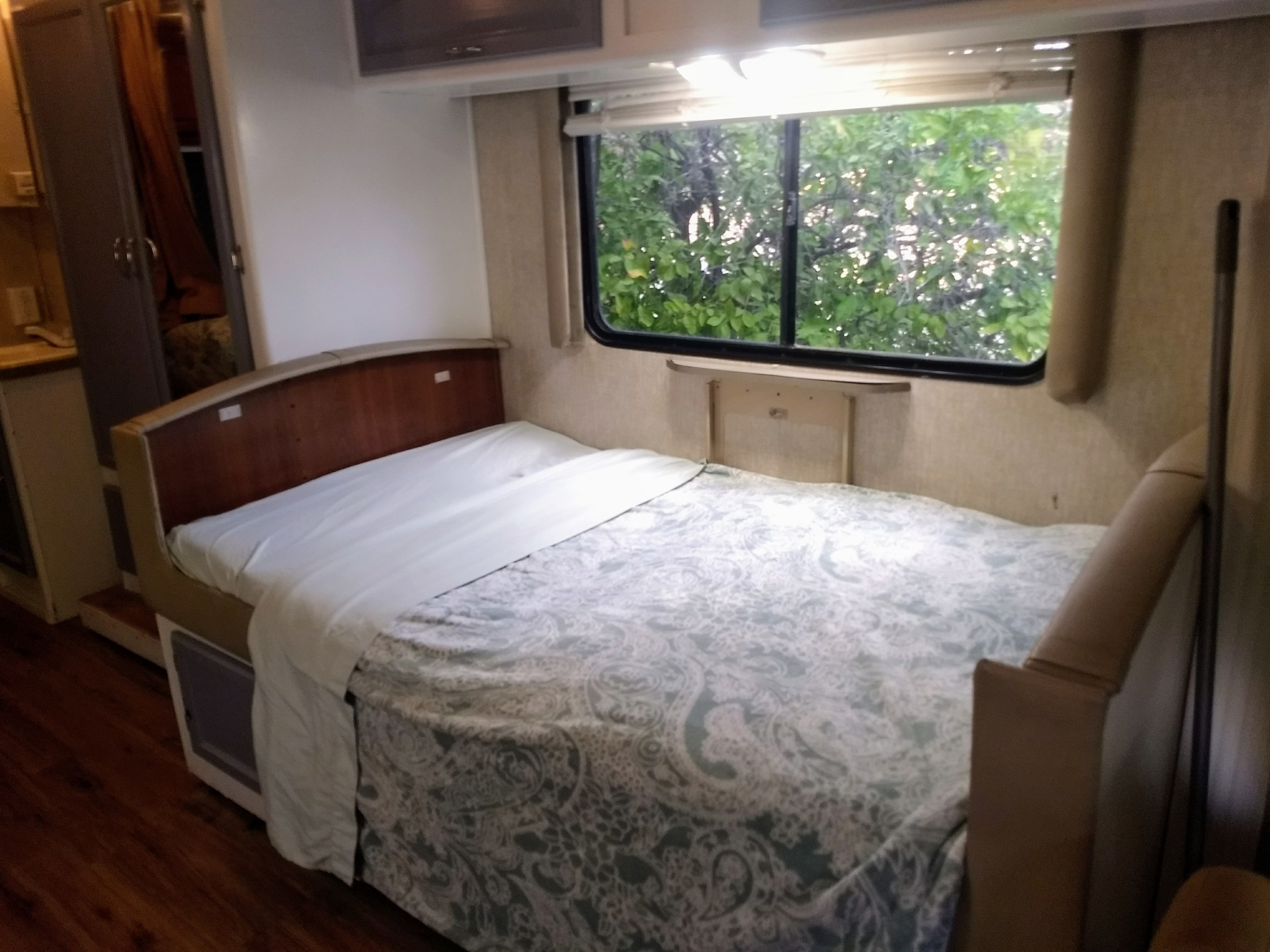 Cozy RV rental bedroom with a neat bed, wooden headboard, and a bright window view of lush green trees.. Fleetwood Jamboree 2009