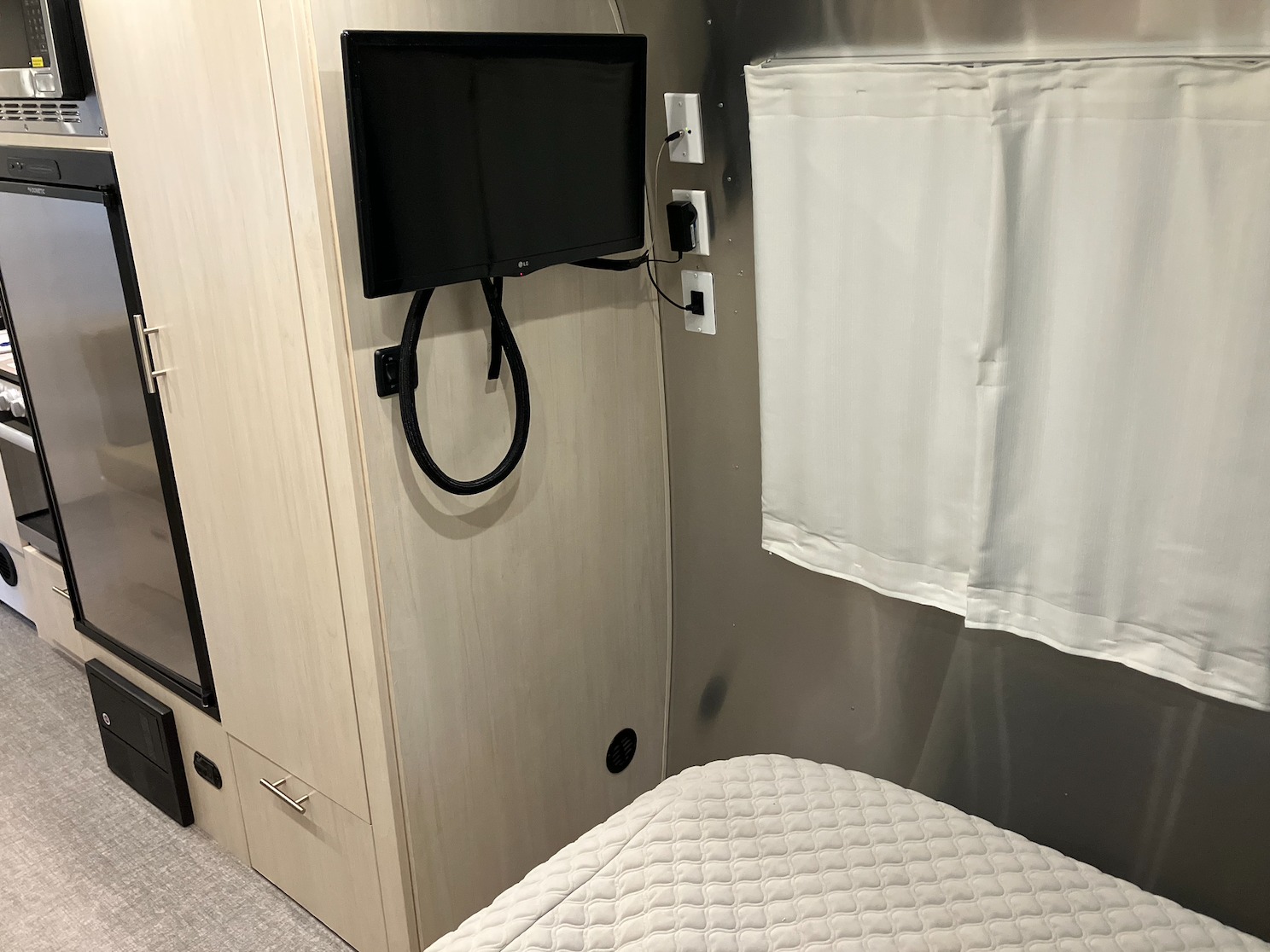 An rv rental bedroom view: a mounted TV, white quilted bed, and light wood cabinetry with a visible fridge.. Airstream Flying Cloud 2021