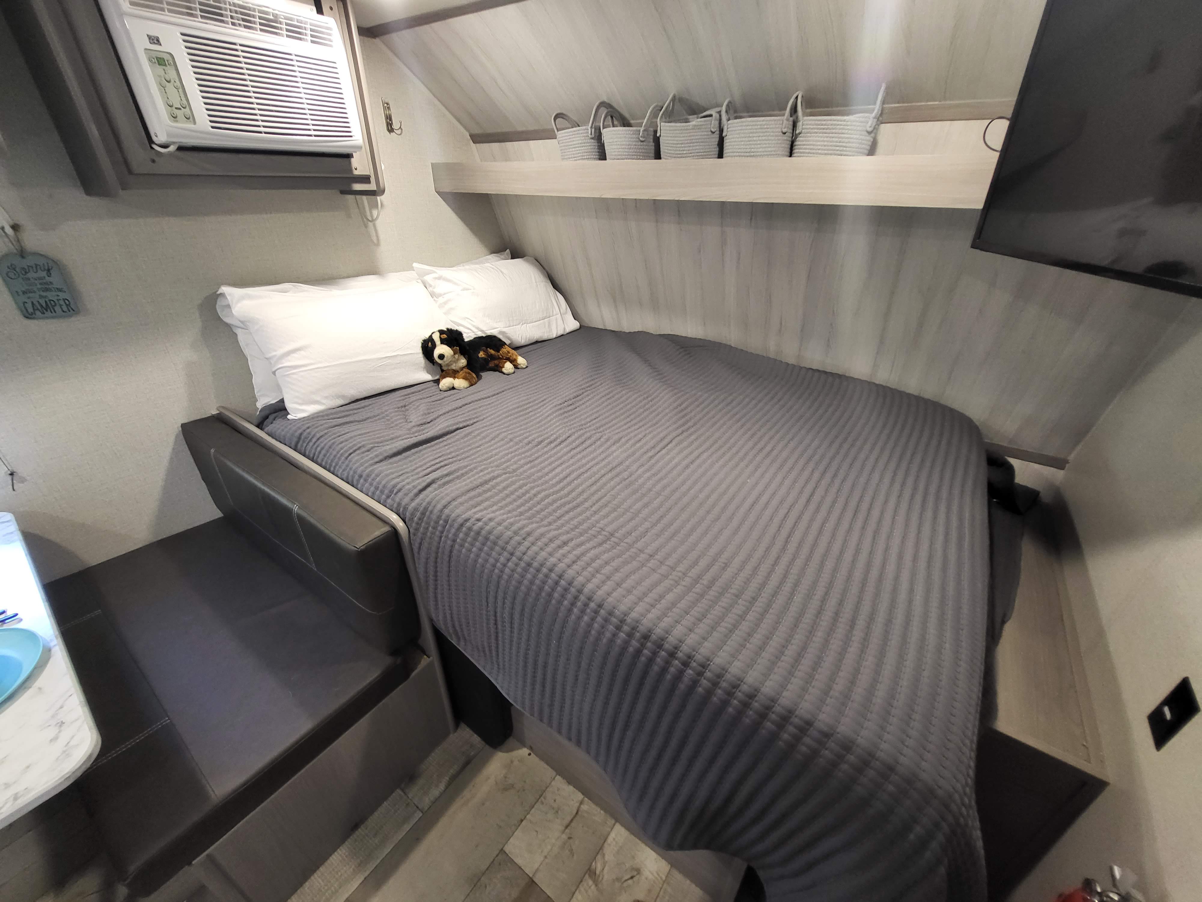 Cozy RV rental interior featuring a bed with a stuffed dog, AC unit, overhead shelf with baskets, and a wall-mounted TV.. Dutchmen Coleman Lantern LT 2023