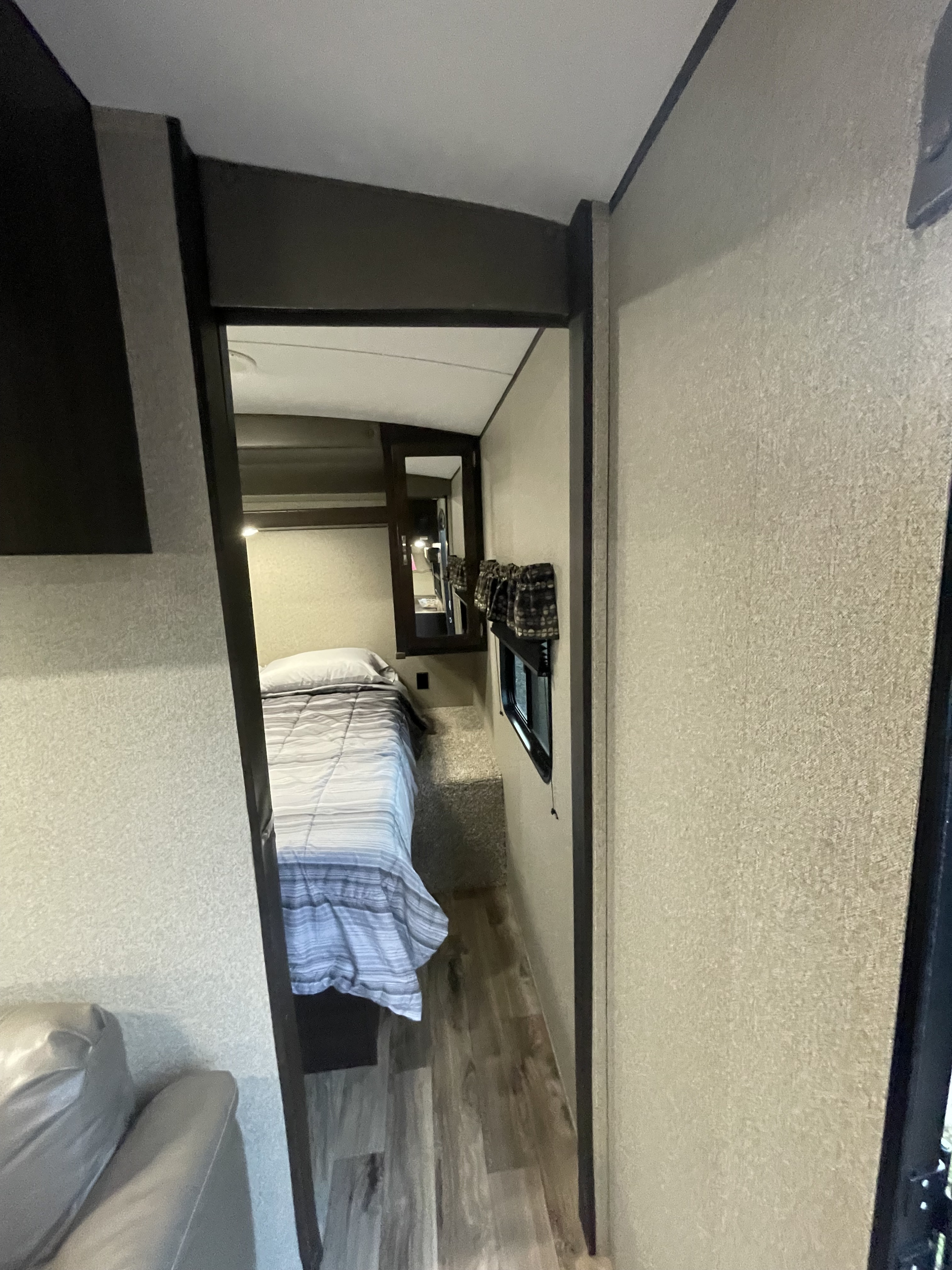 RV rental's compact bedroom with striped bedding, mirror, and window, seen through a narrow hallway from the living space.. Keystone RV Springdale 2020