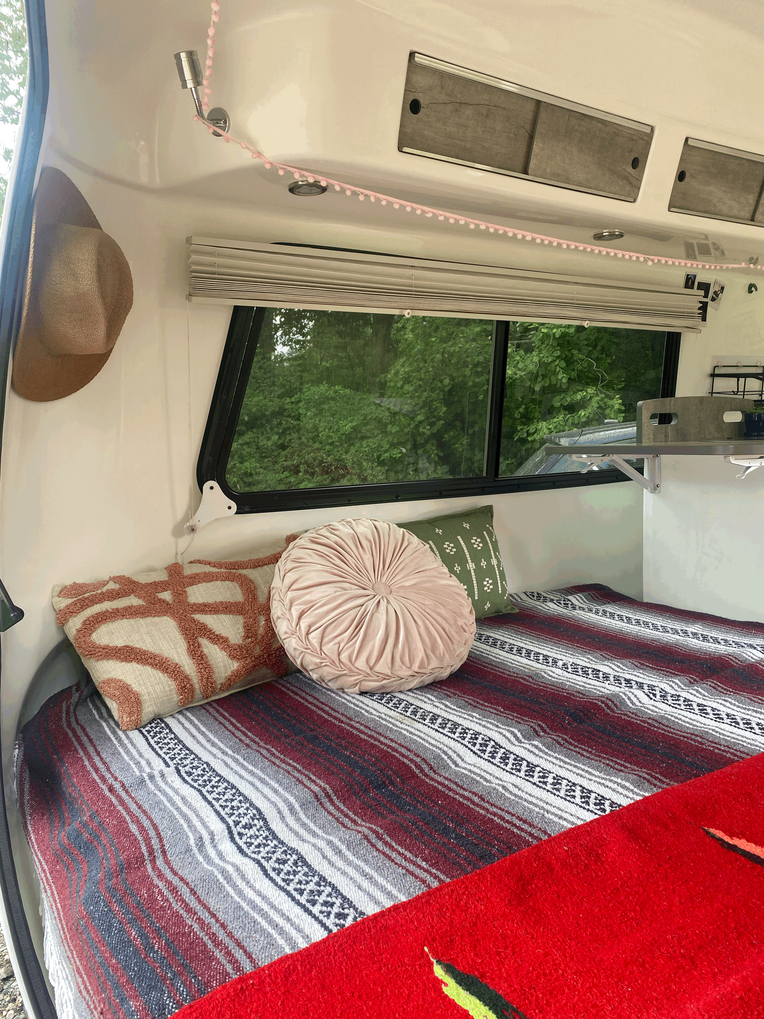 Cozy RV rental interior with a comfortable bed, colorful blankets, decorative pillows, and a window view of lush trees.. Happier Camper HC1 2022