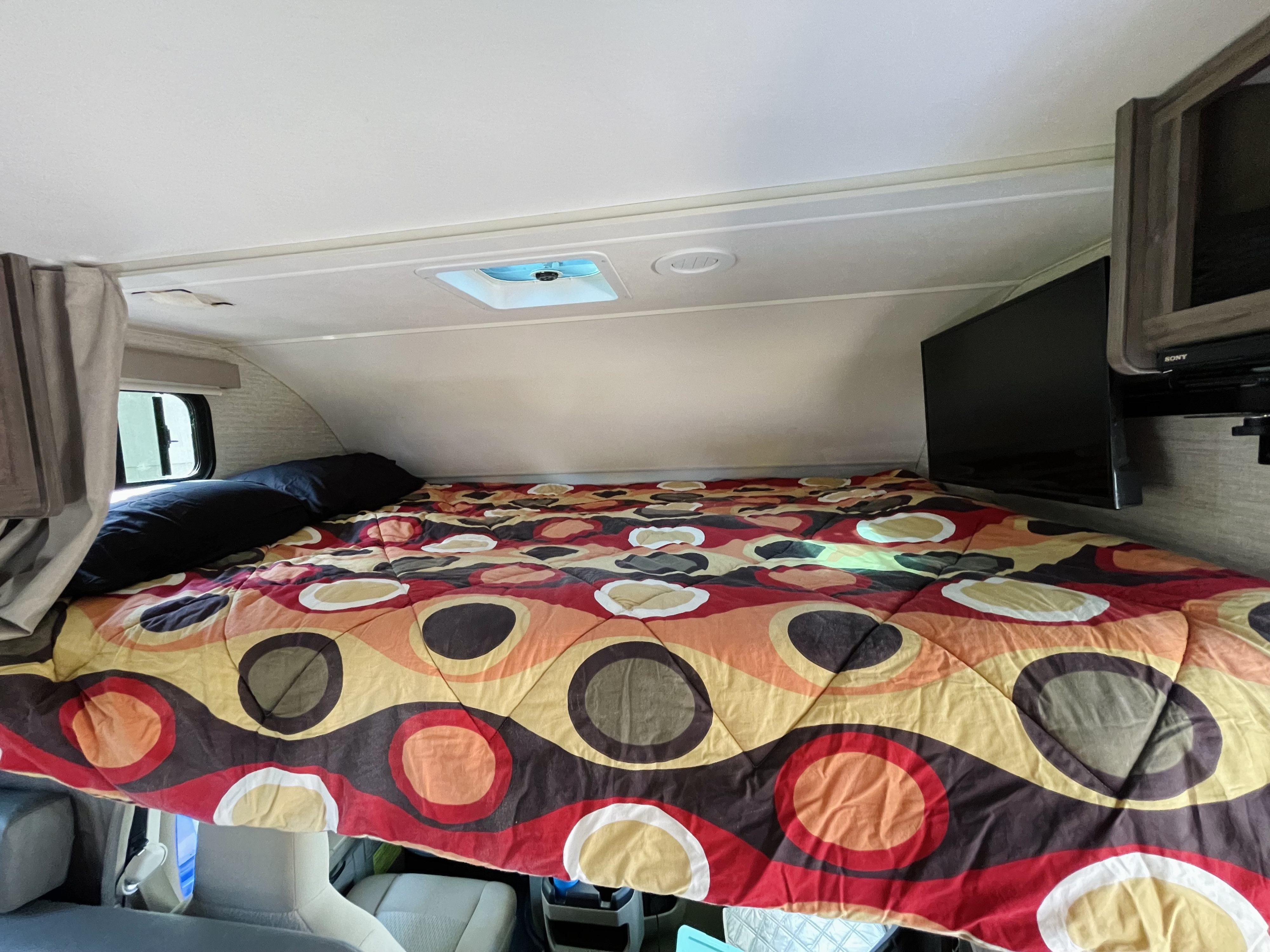 Cozy cab-over bed in an RV rental, featuring a vibrant comforter, window, and mounted TV. Perfect for road trips.. Winnebago Minnie Winnie 2020