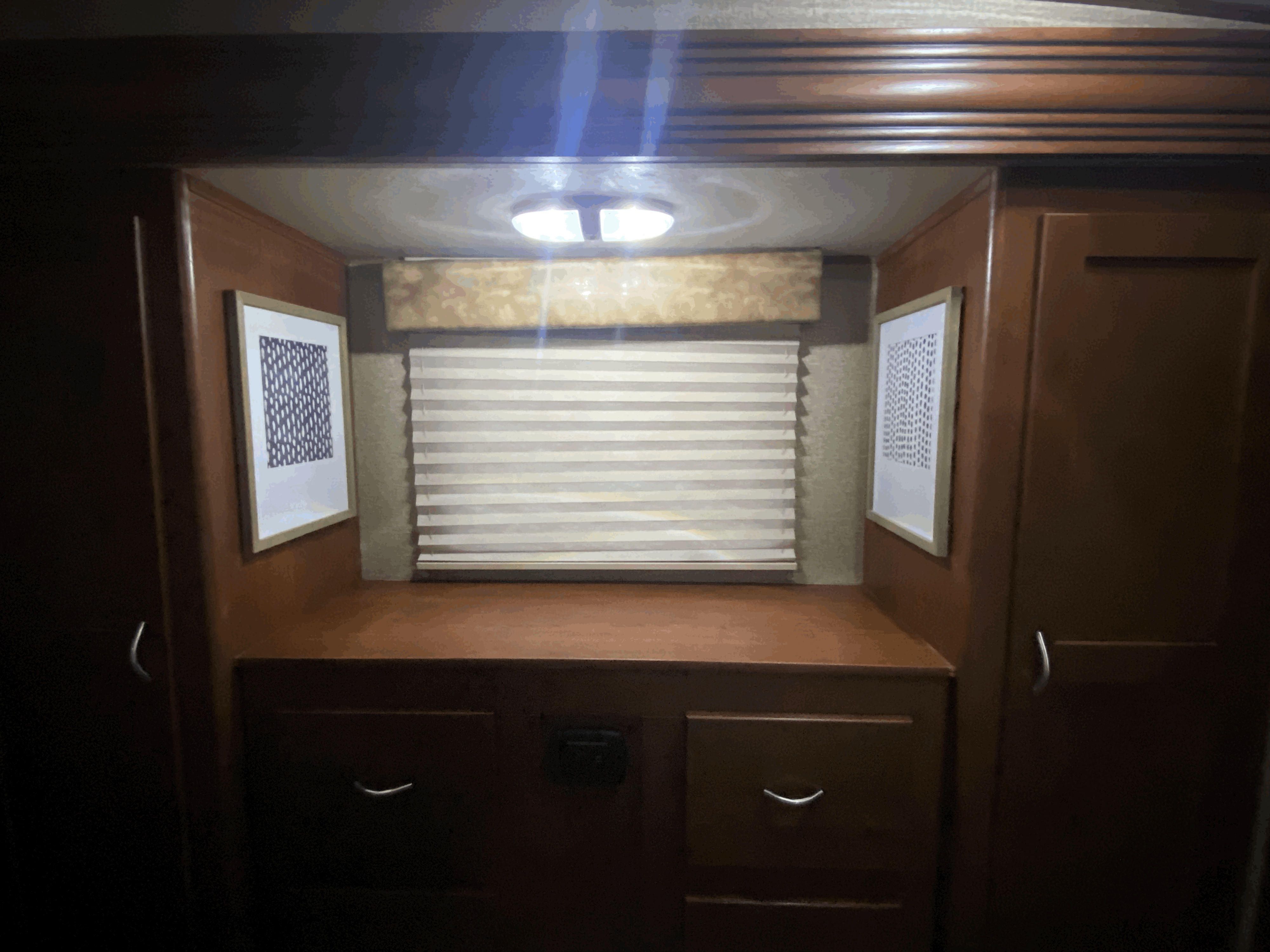 Cozy RV rental bedroom vanity with rich wood cabinets, a window with blinds, framed art, and overhead lighting.. Forest River Vengeance 2015