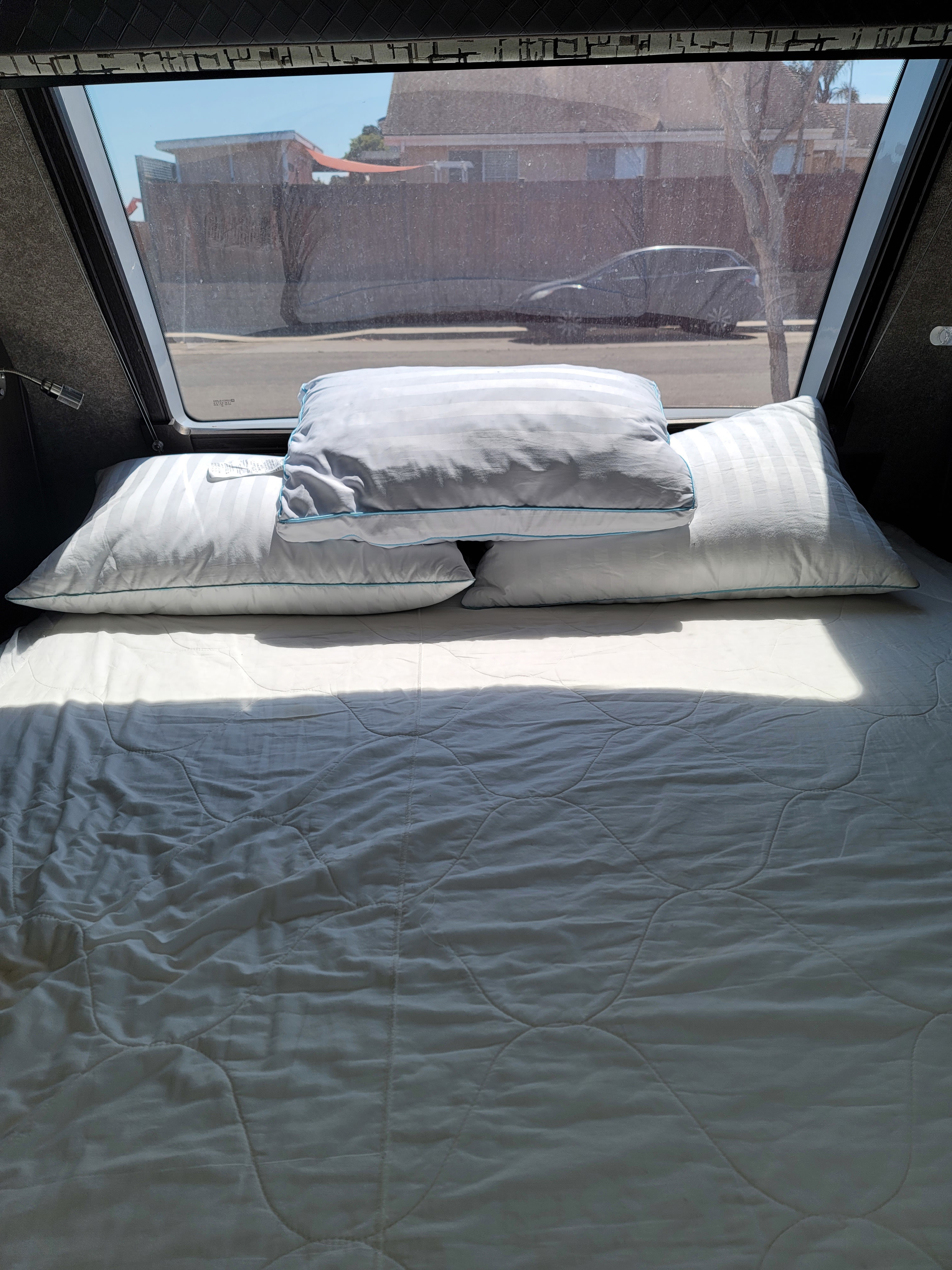 Cozy bed inside an RV rental. Plump pillows await, with a view of a residential street and parked car through the window.. Jayco White Hawk 2019