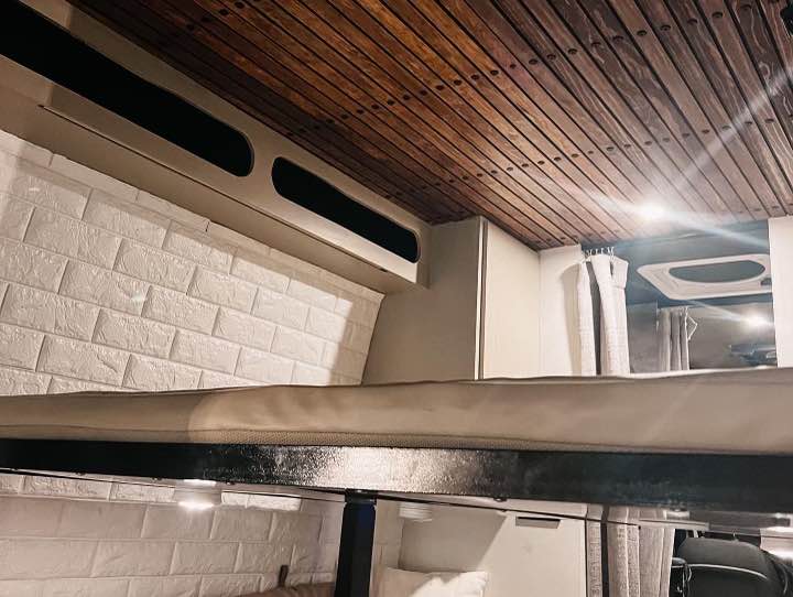Explore this RV rental's cozy interior, featuring a wooden slatted ceiling, a bunk bed, and stylish white brick-patterned walls.. Mercedes-Benz Sprinter 2019