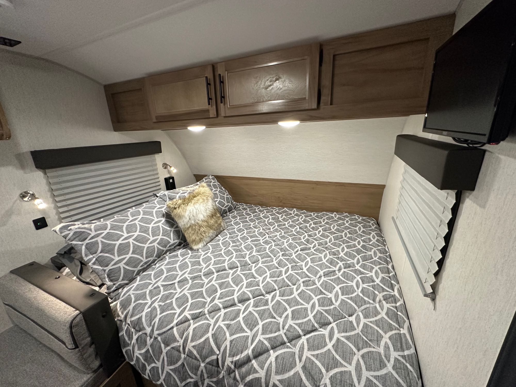 Compact RV rental bedroom with patterned bed, overhead cabinets, reading lights, window blinds, and TV.. Forest River Wolf Pup 16 BHS Black Label 2023
