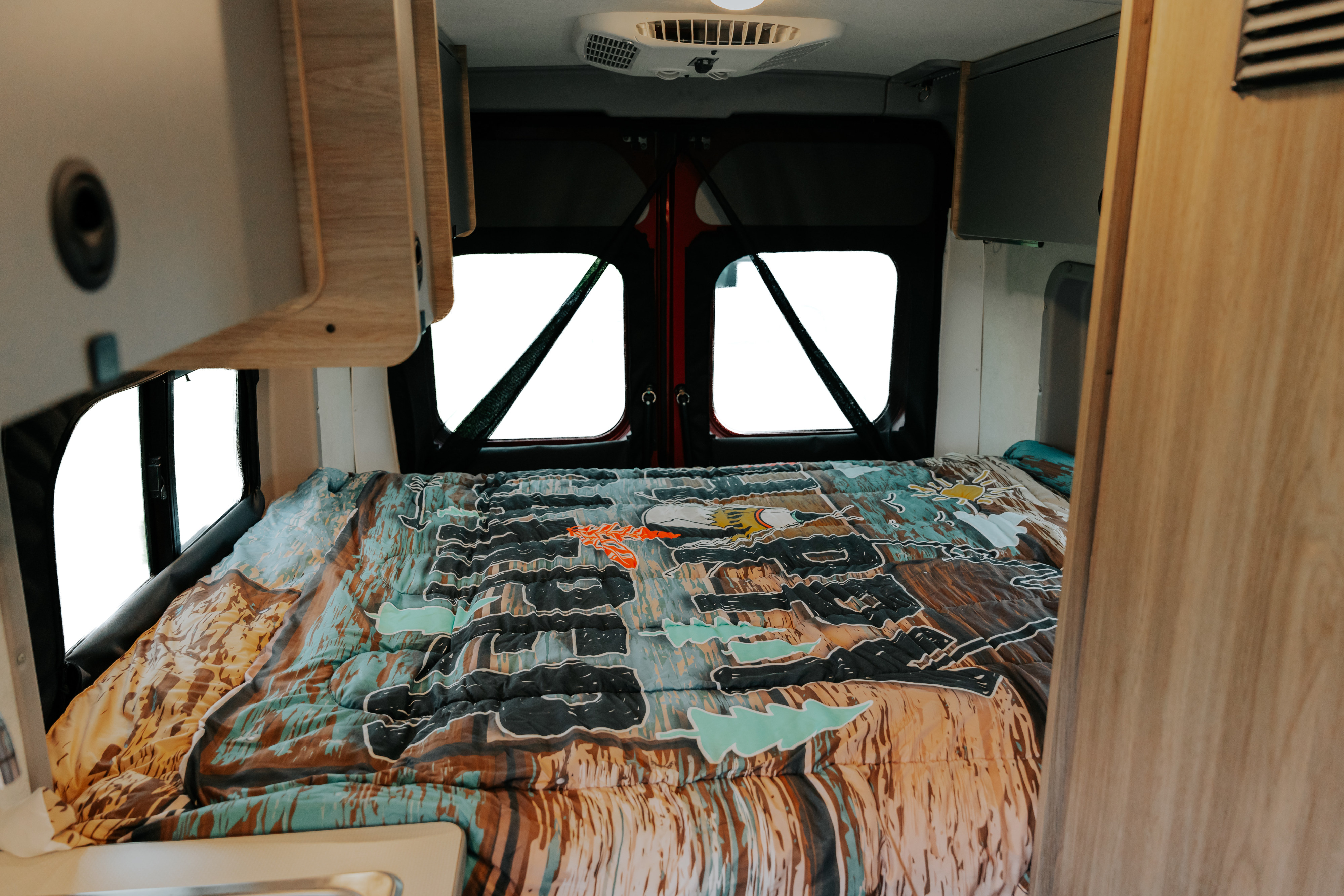 RV rental interior: a comfortable bed with a colorful patterned comforter, visible bright windows at the back.. Winnebago Winnebago 2023