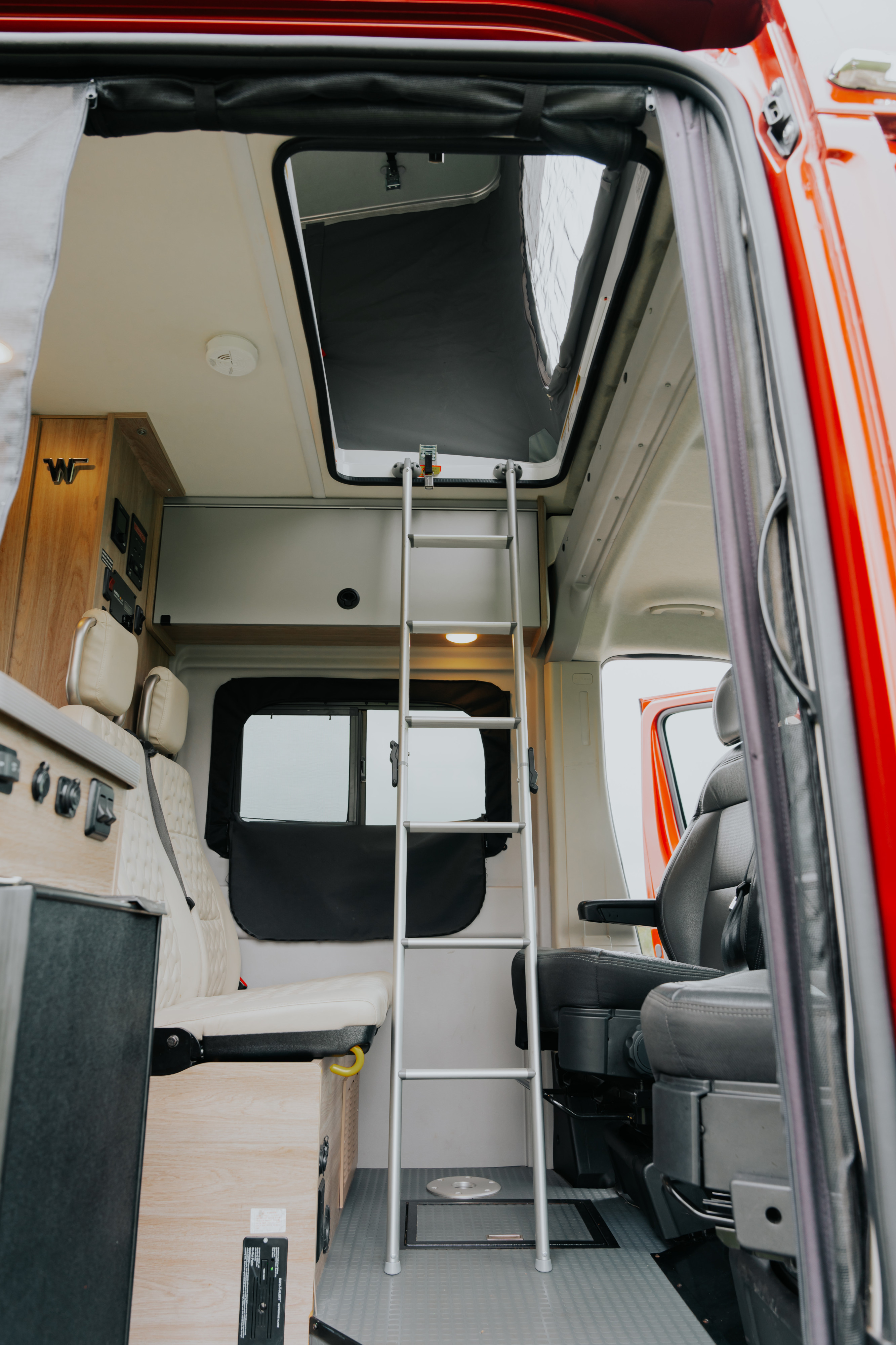 Inside an RV rental: a ladder to a pop-top bed, plush seating, and wooden cabinetry with control panels.. Winnebago Winnebago 2023