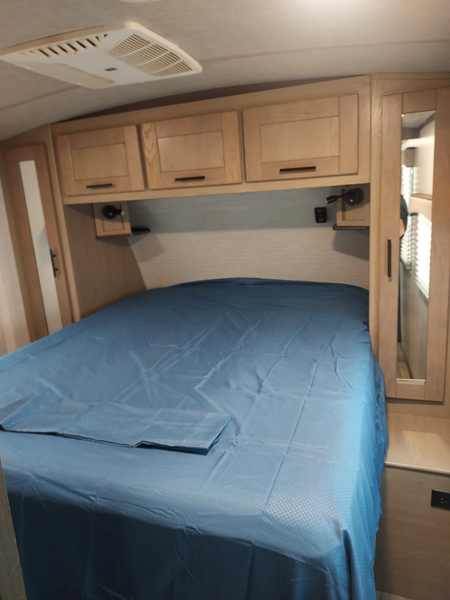 RV rental bedroom featuring a bed with blue cover, light wood overhead cabinets, and a ceiling AC unit.. Heartland RVs Sundance Ultra Lite 2020