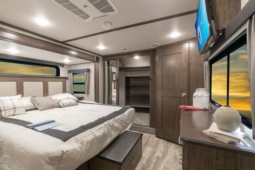 Spacious RV rental bedroom with a large bed, wall-mounted TV, and window showcasing a golden sunset view.. Grand Design Solitude 2020