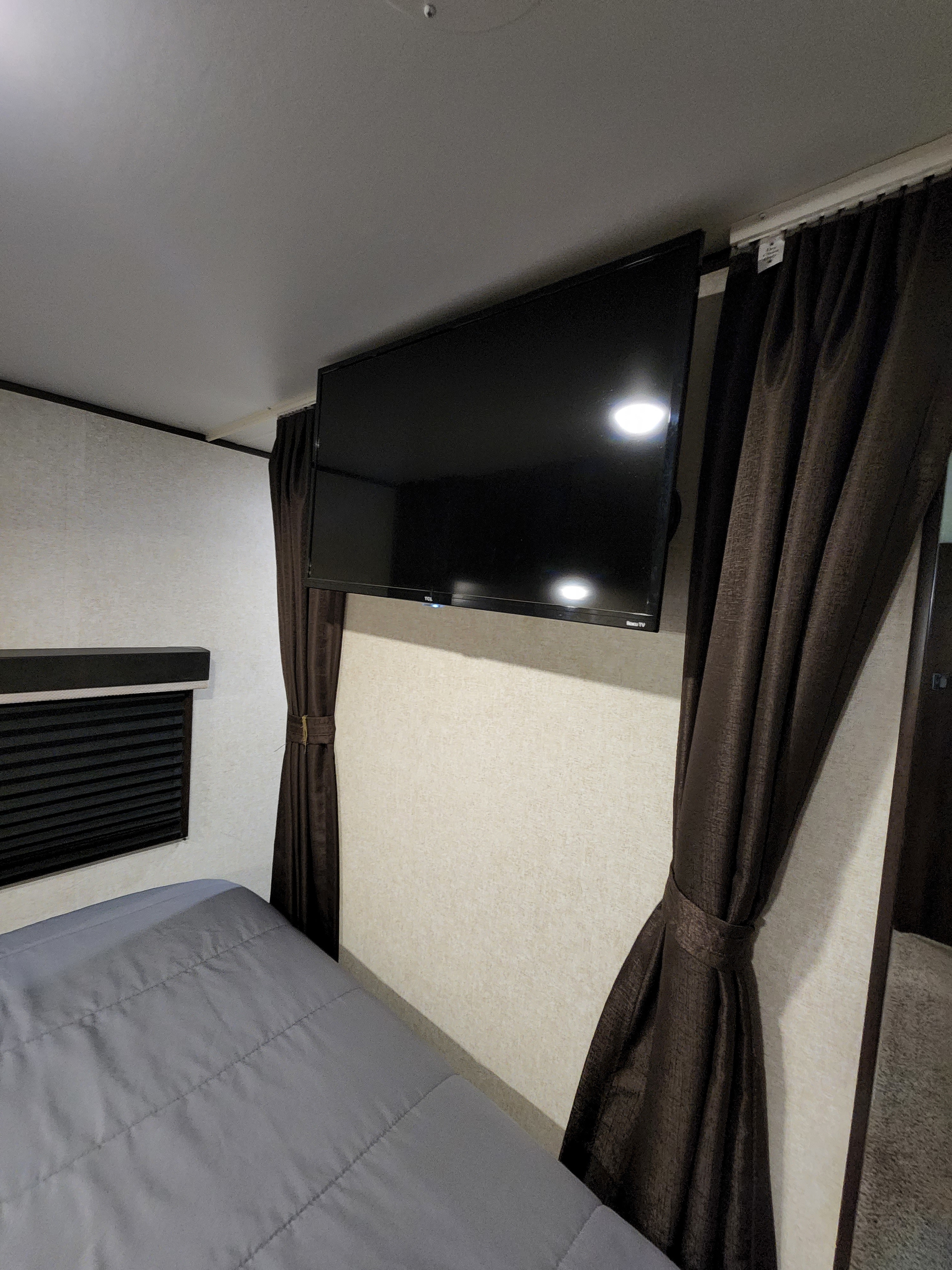 Cozy RV rental bedroom features a mounted TV, comfortable bed with gray comforter, and tied-back curtains.. Jayco Jay Flight SLX 2019